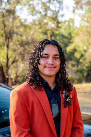 A person in a bright orange suit jacket with curly hair stands for a formal photo outdoors.
