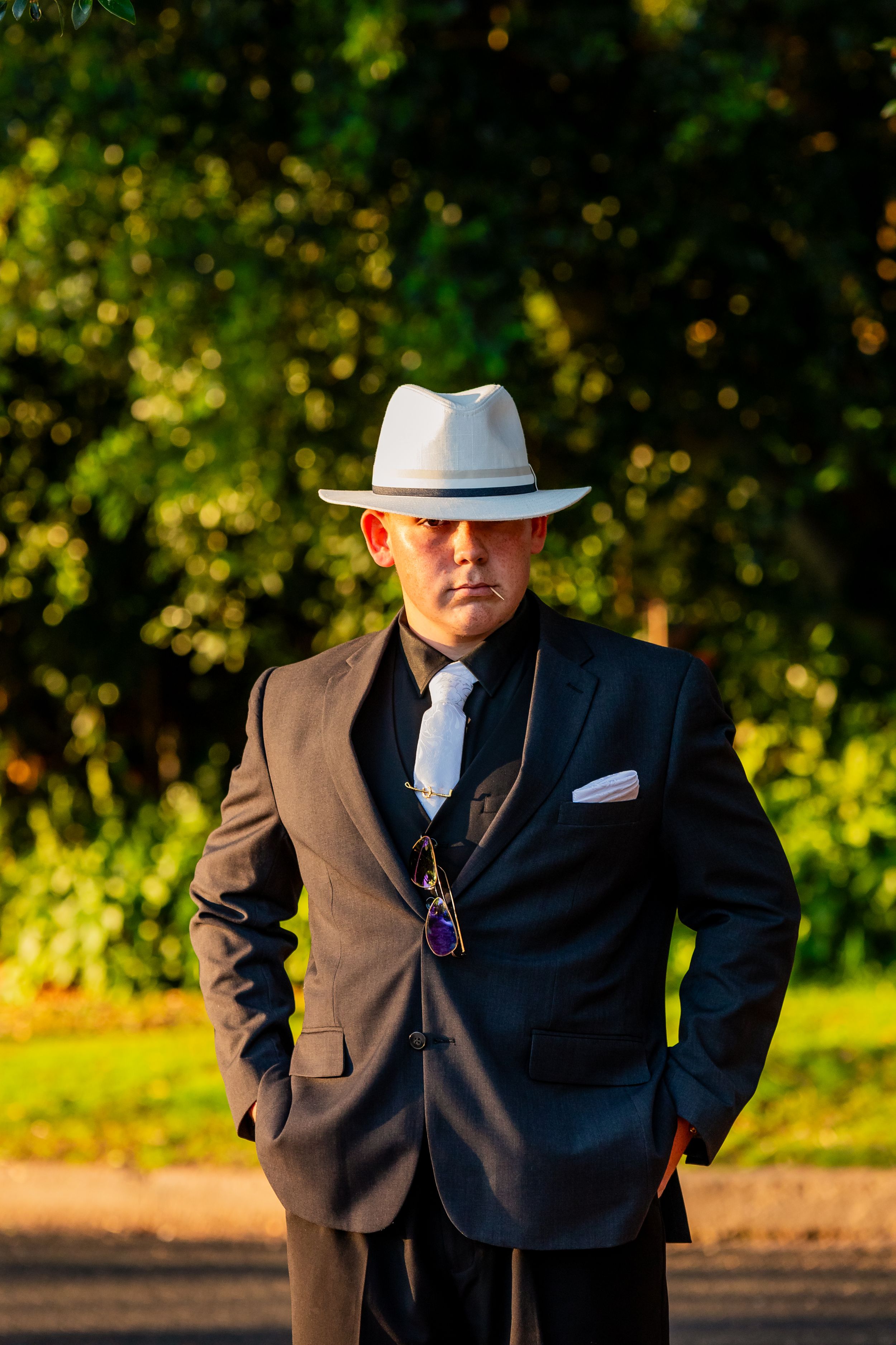 A smartly dressed figure in a dark suit and white fedora poses in front of lush greenery.