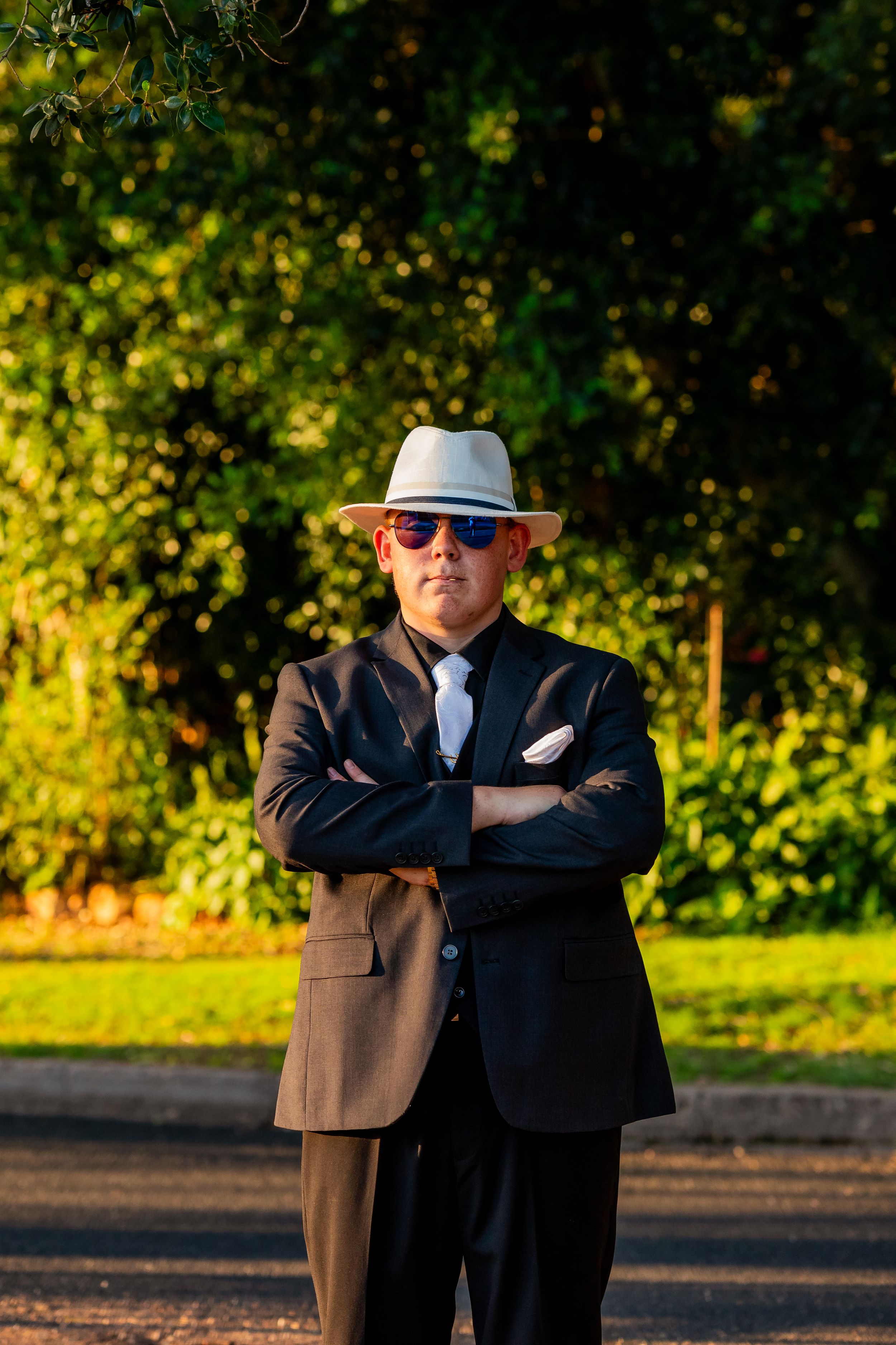 A formal figure in a black suit and fedora strikes a confident pose against a sunlit garden backdrop.