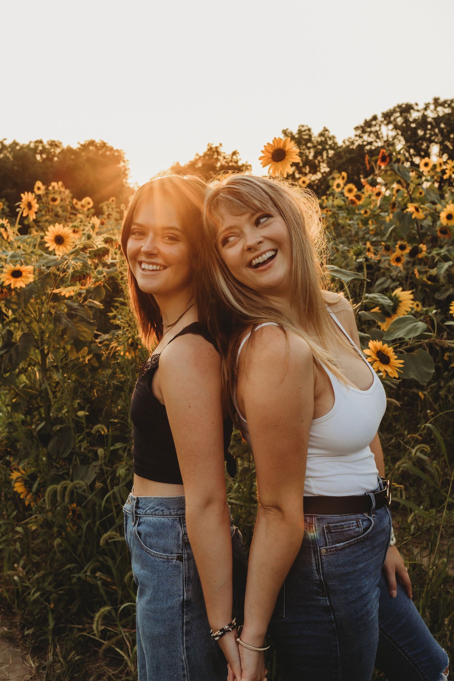 Two friends laugh together in a sunflower field at golden hour while wearing casual summer outfits.