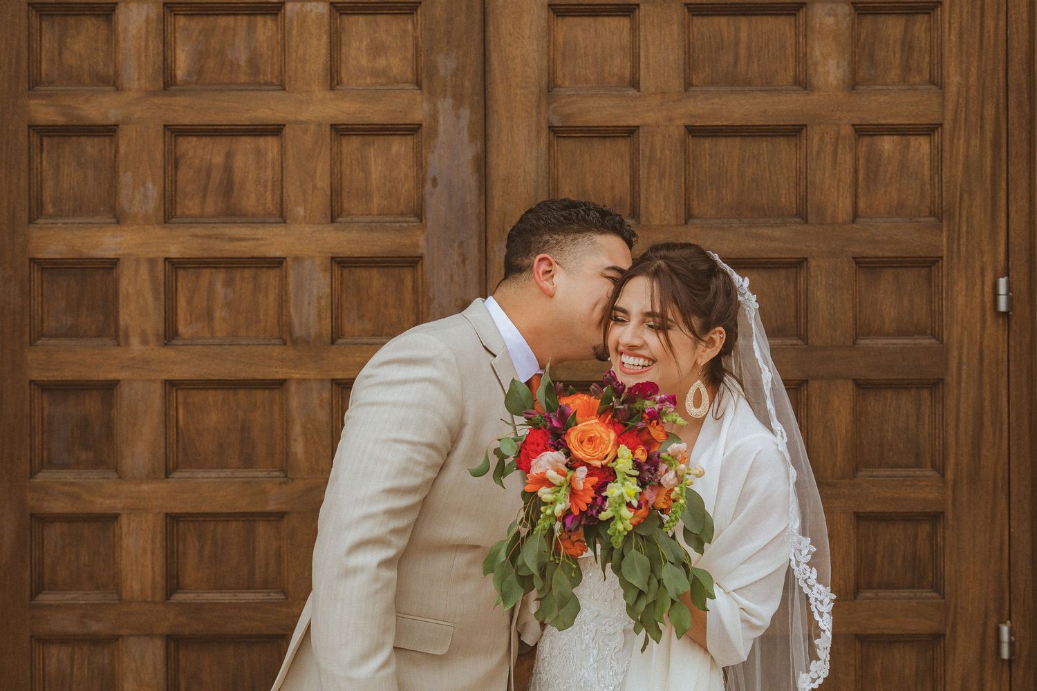 Newlyweds sharing intimate moment with orange and red bouquet against wooden door.