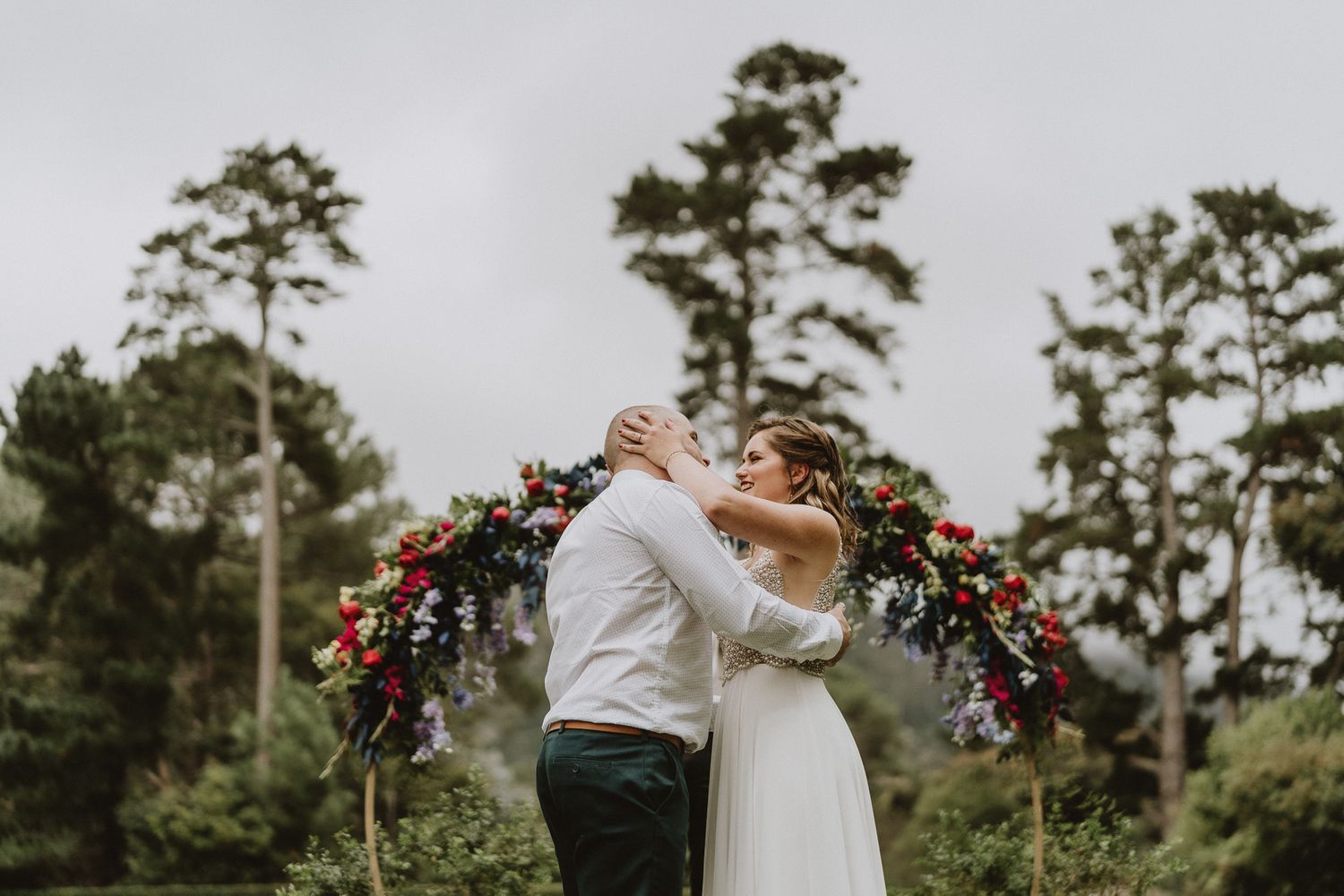 Newlyweds embrace under a floral arch with tall pine trees in the background.