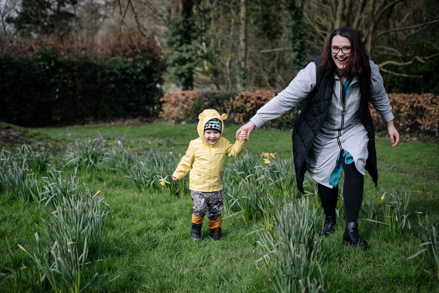 A child in a yellow raincoat running through spring flowers with adult companion.