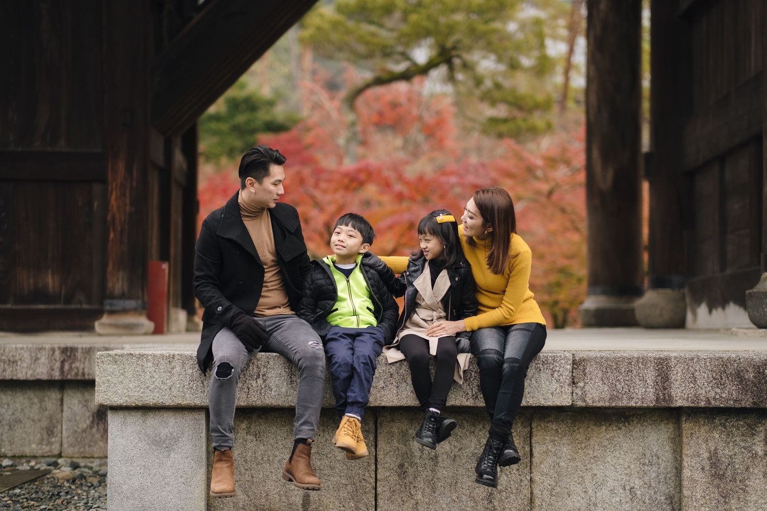 Family of four sitting on stone wall laughing together with colorful autumn foliage in Japanese temple.