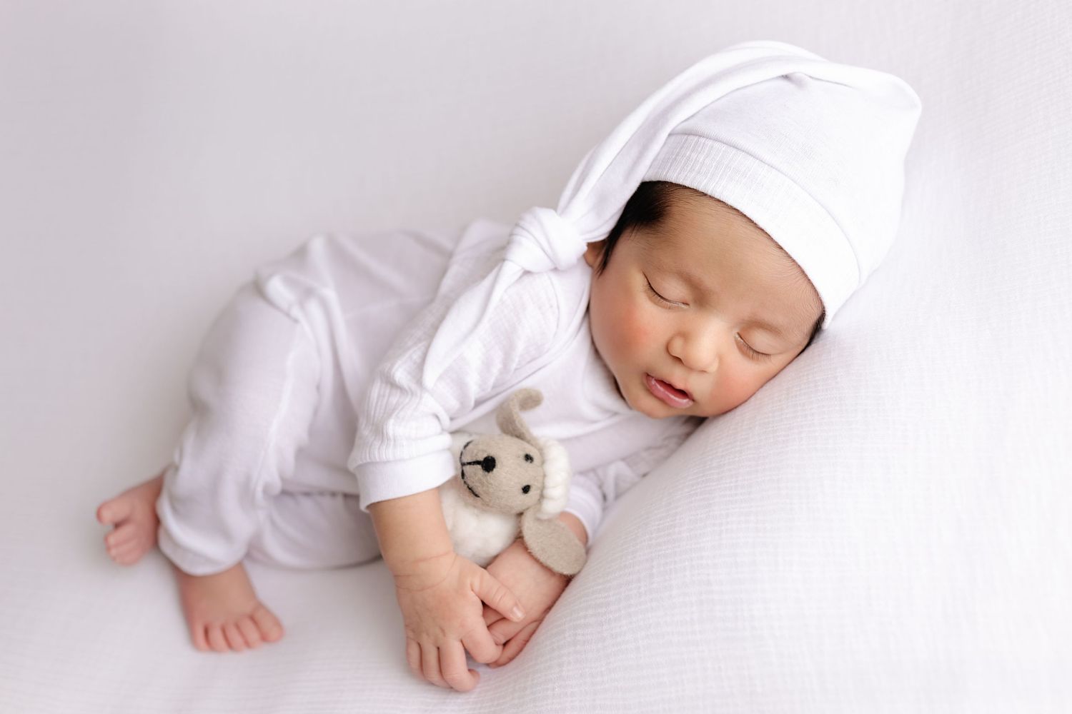 Sleeping baby in white clothing and hat cuddles small teddy bear while lying on soft white surface.