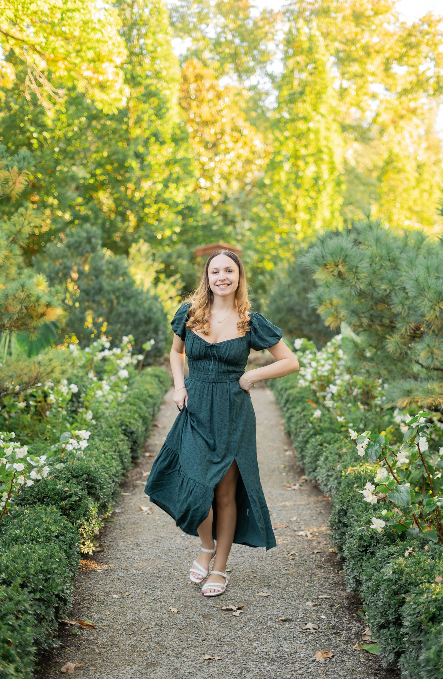 A young woman in a dark green midi dress walks along a garden path lined with white flowers and shrubs.