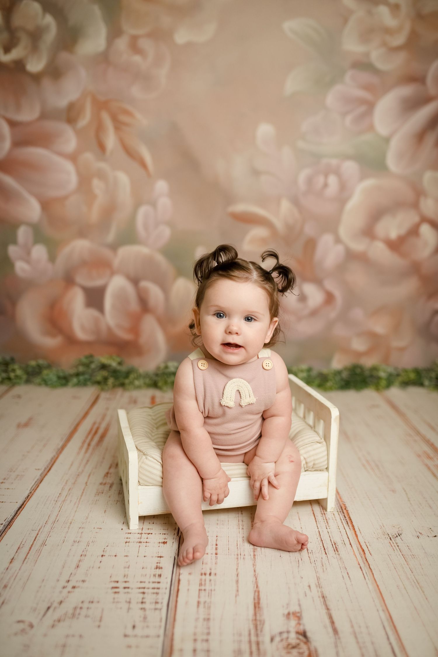 Professional baby photo session with vintage white bed against floral backdrop.