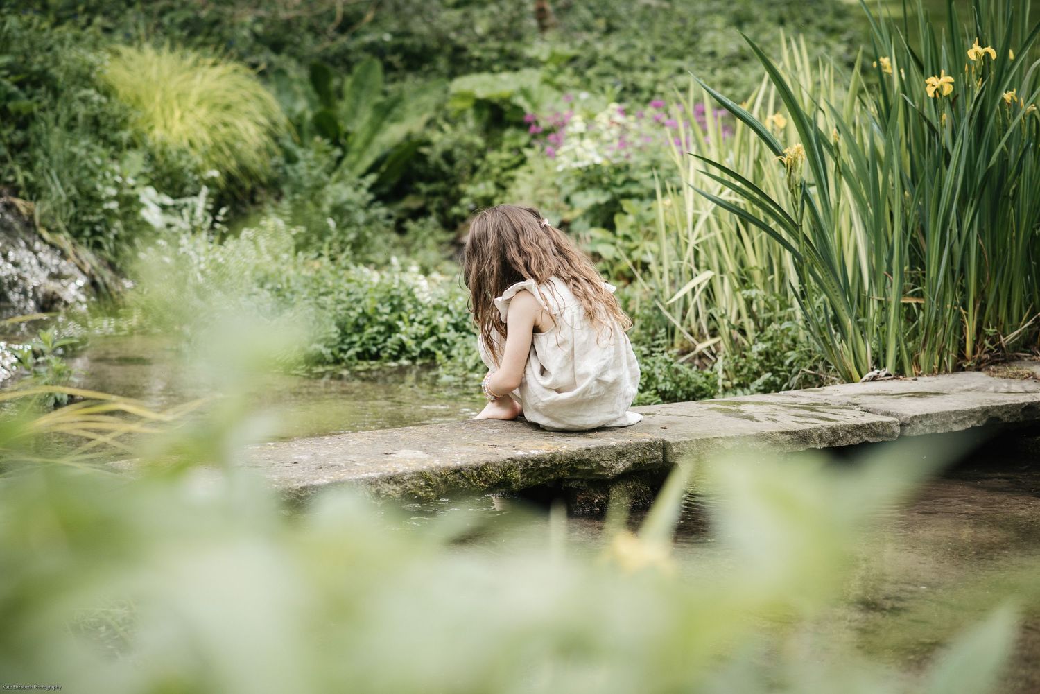 A person sits contemplatively by a pond surrounded by lush garden greenery.