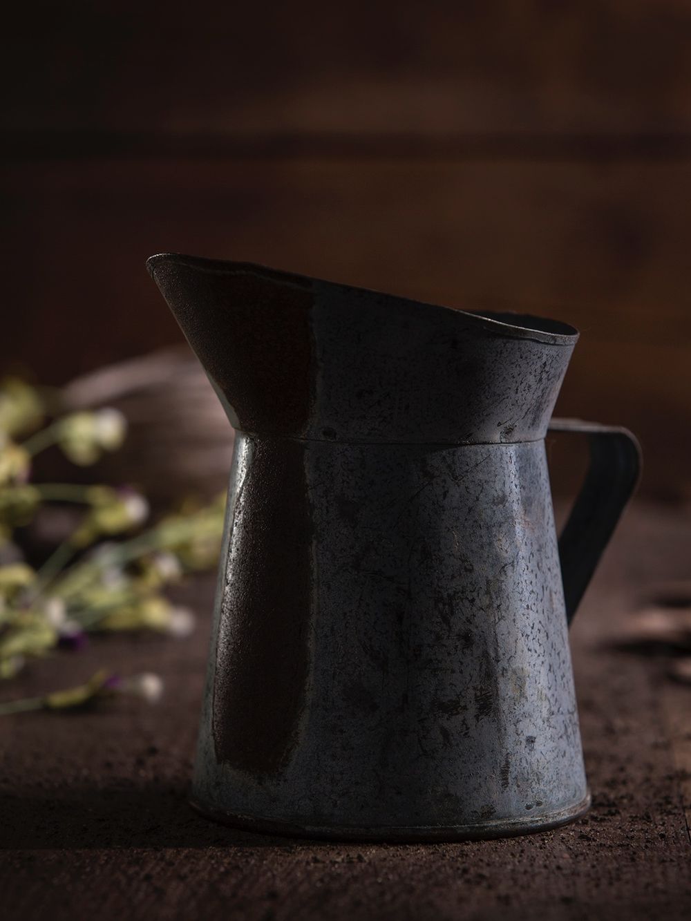 A vintage tin water pitcher sitting in front of fresh cut flower buds and a bundle of twine with dark and moody lighting