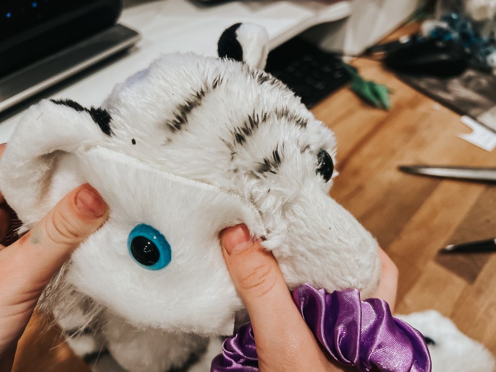 A hand holds ups a piece of faux white fur with a blue doll eye in it to a damaged stuffed white tiger as she repairs it