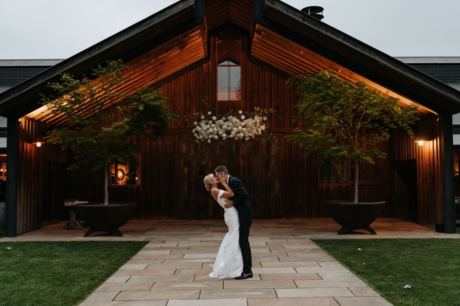 Bride and groom share a romantic dip kiss outside a rustic dark wood barn venue at dusk.