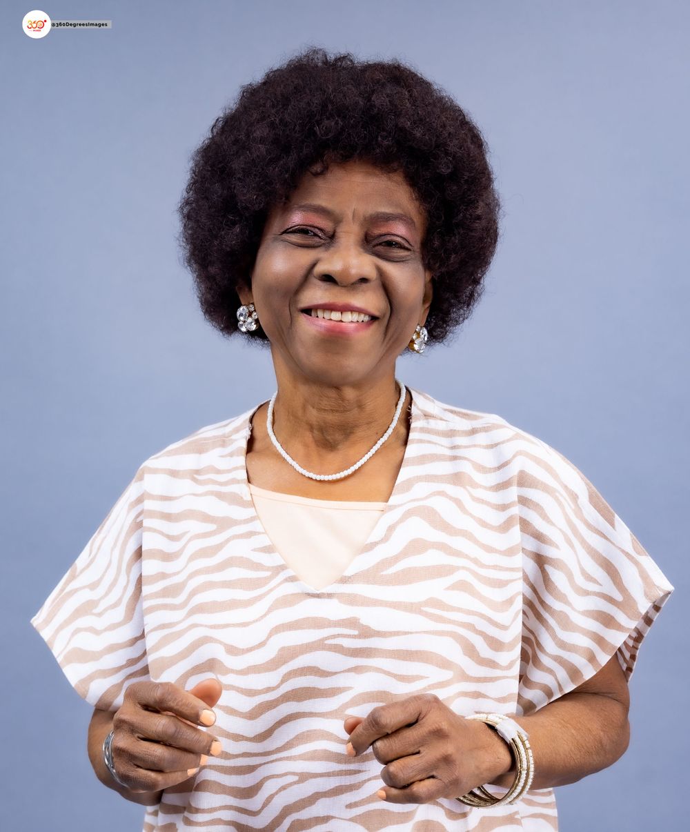 Smiling elderly Black woman with afro hair wearing a zebra-print blouse and pearl necklace against a blue background.