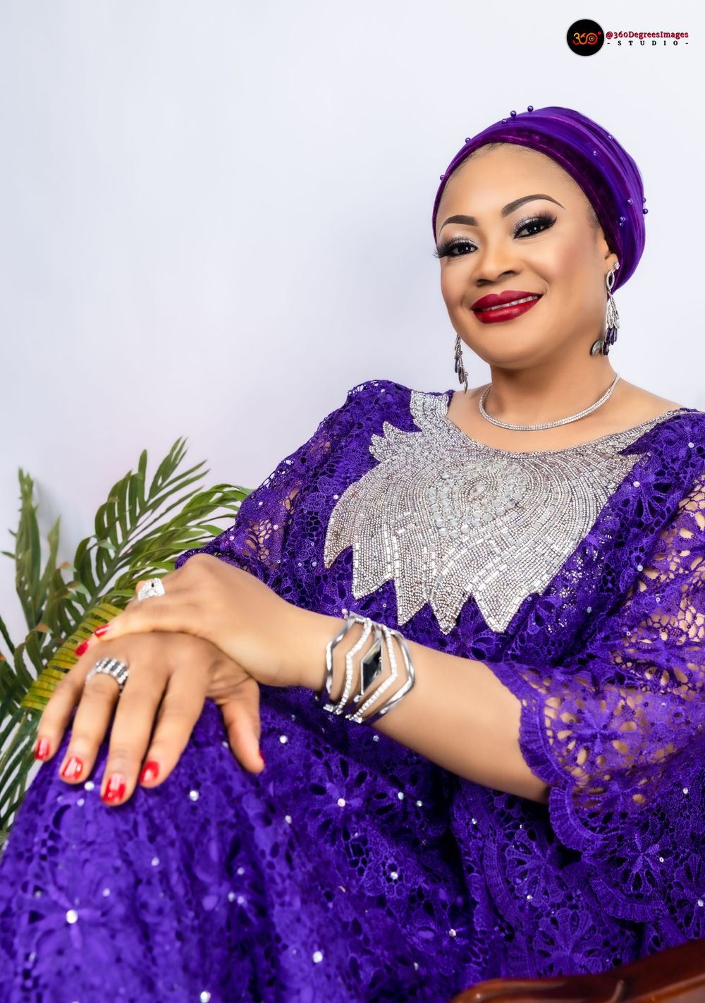 A smiling woman in a purple sequined lace boubou with silver embroidery and matching headwrap poses seated gracefully.