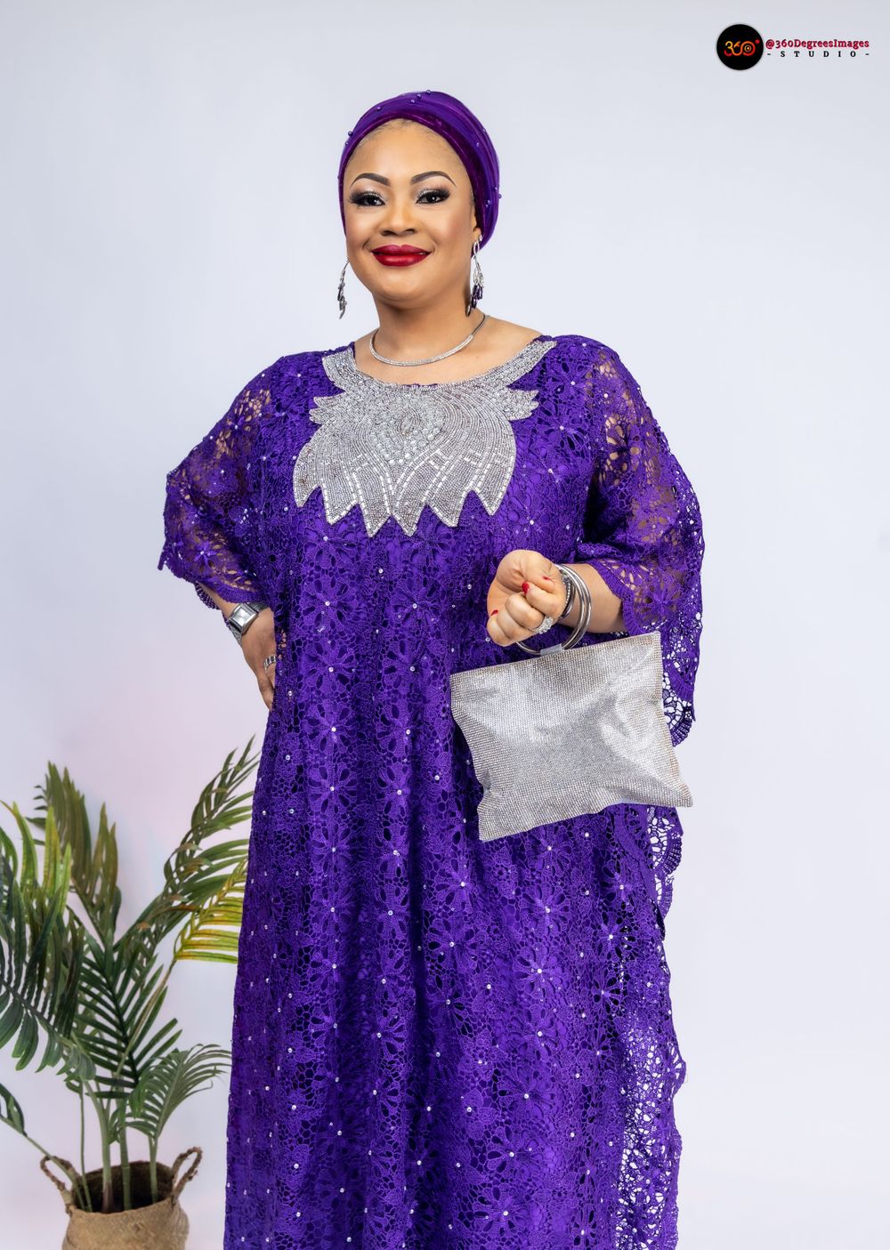 A woman poses in an elegant purple lace sequined boubou dress with silver embroidery and matching headwrap holding a clutch.