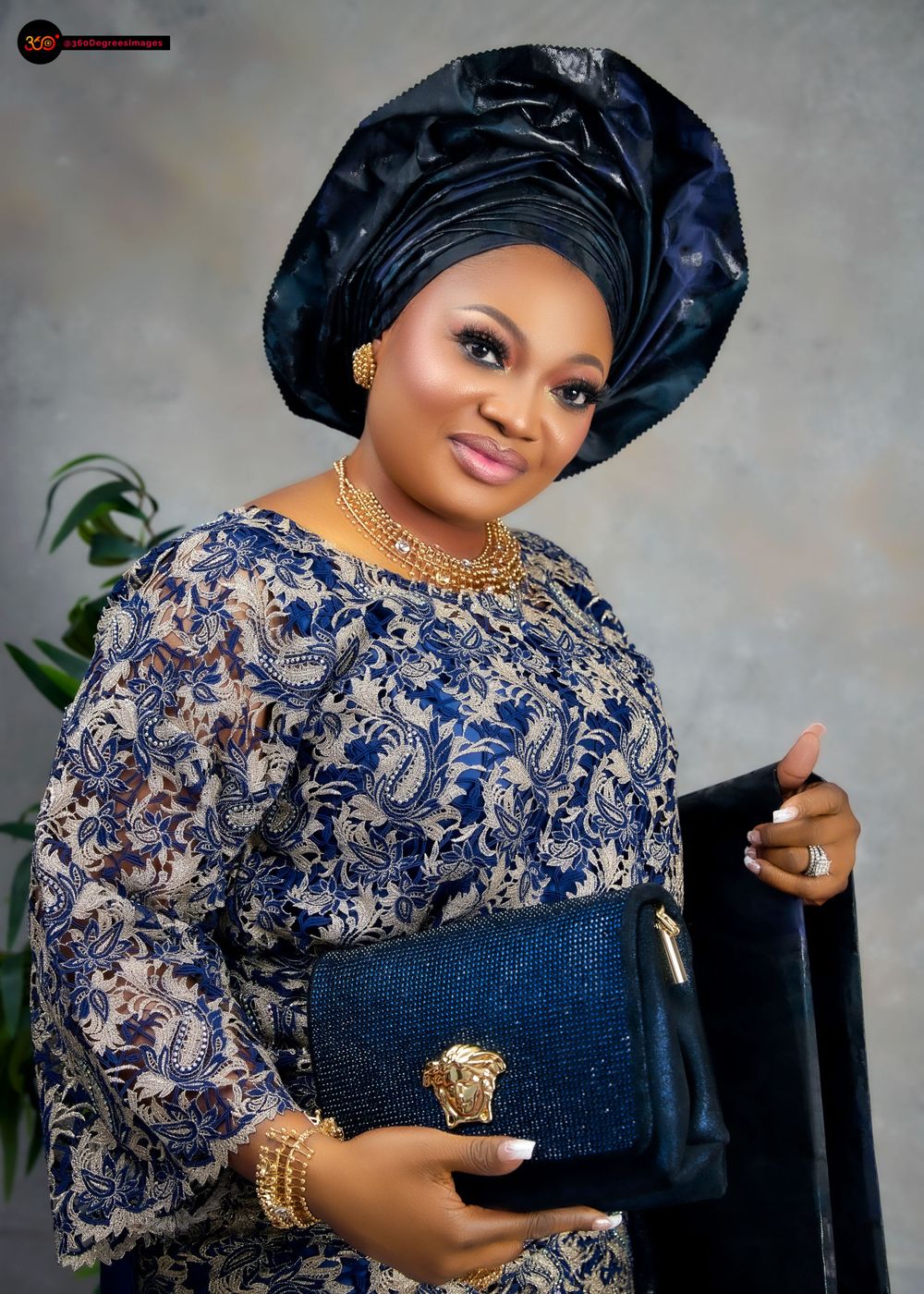 Woman in navy blue embroidered Nigerian outfit with dark gele headwrap holds blue Versace clutch bag.
