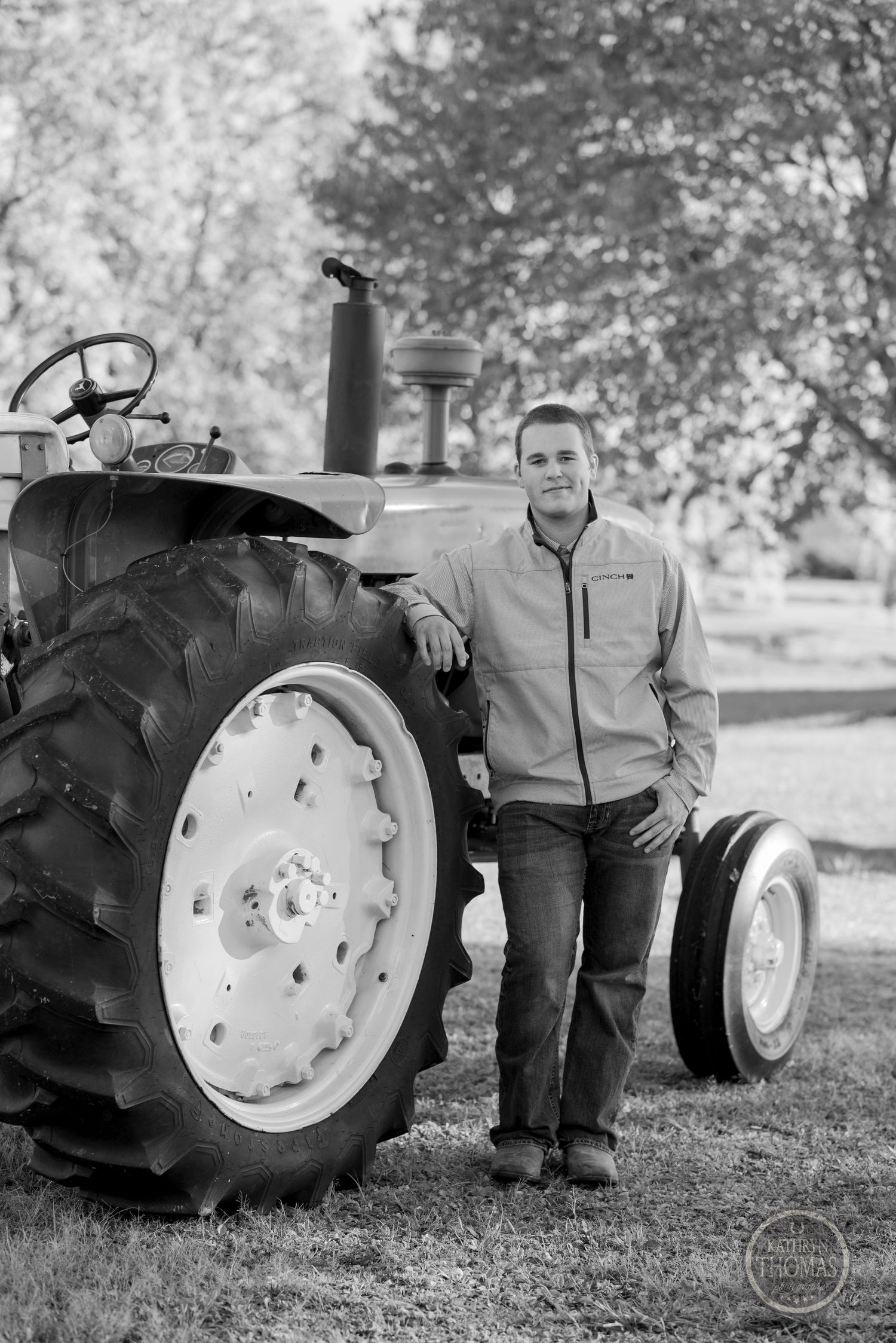 Black and white portrait of person standing beside large vintage tractor wheel.