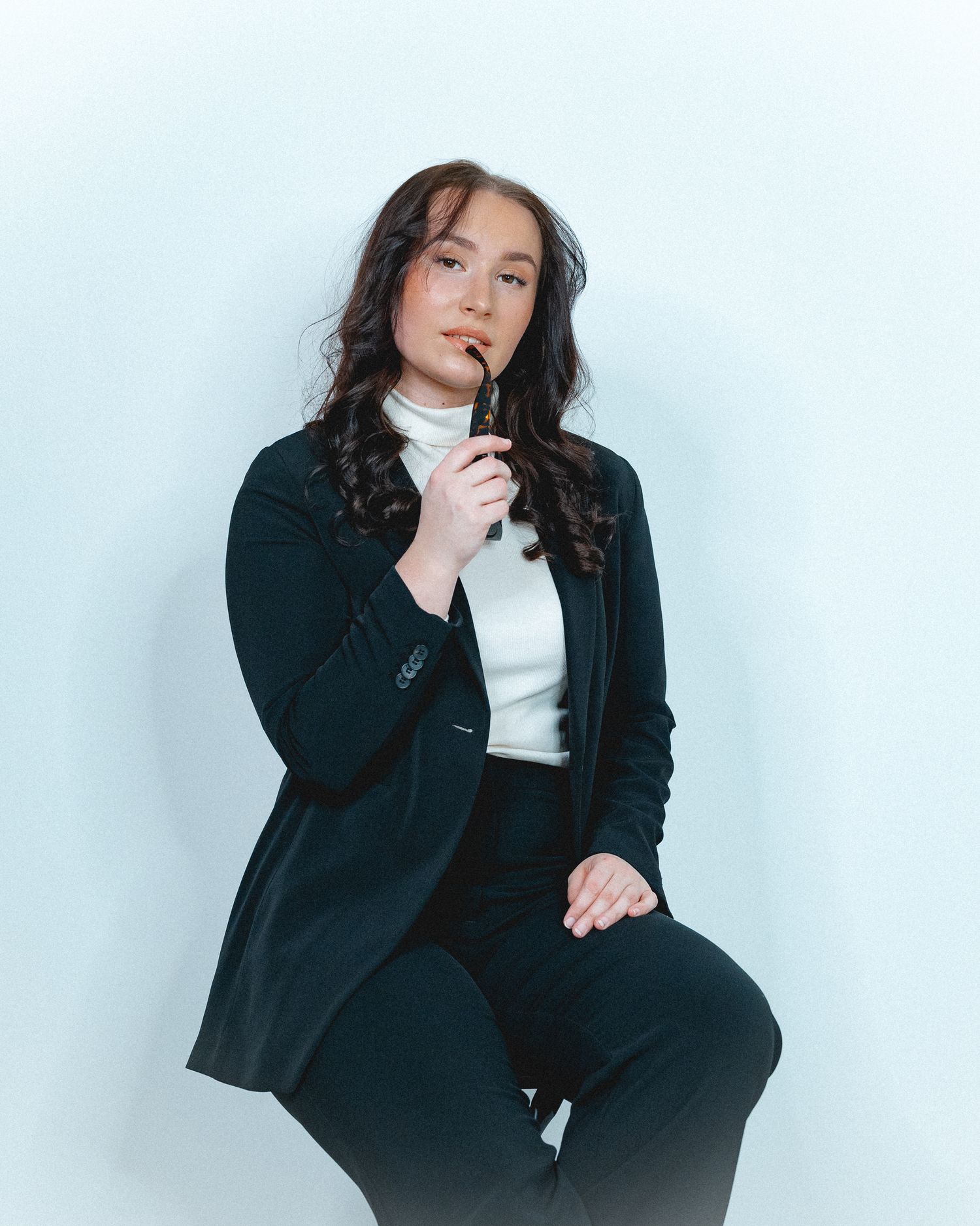 Fashion portrait of a person in black blazer and white turtleneck against light background.