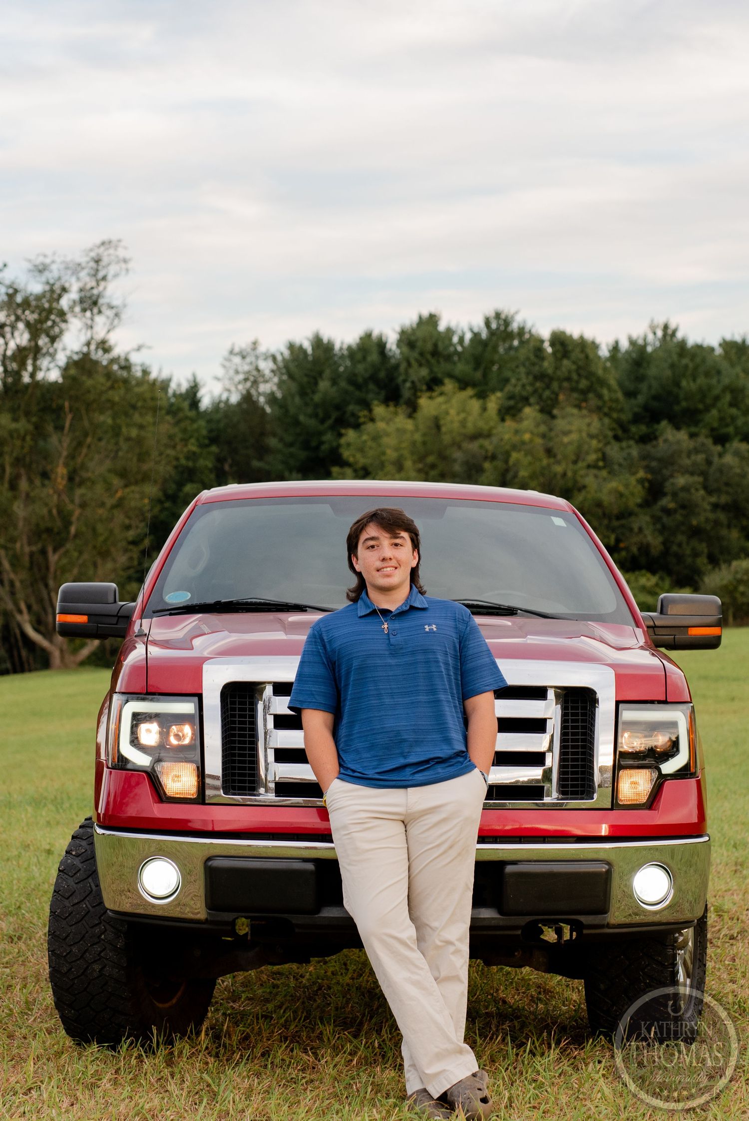 A person in a blue polo stands in front of a red Ford F150 pickup truck in an open field.
