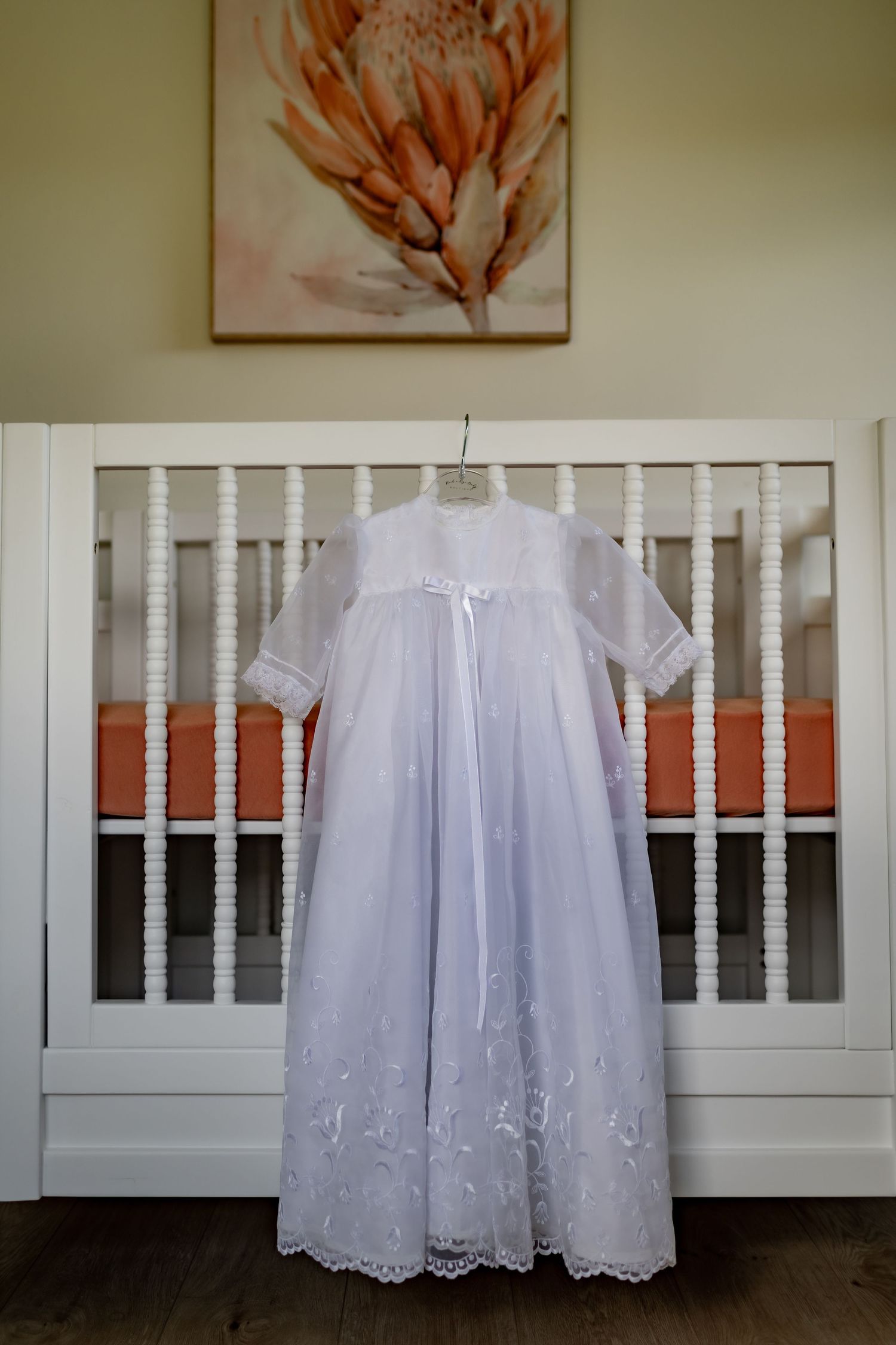 White christening gown with lace details hanging on display against a white crib and floral artwork.