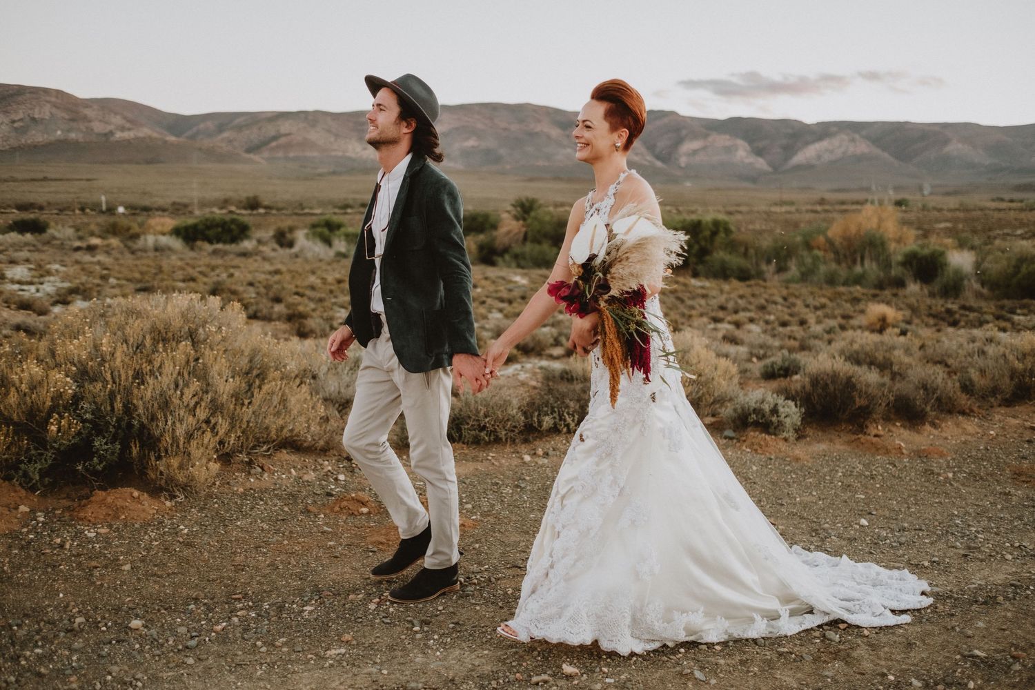 A couple poses in desert terrain with mountains and dramatic clouds in the background.