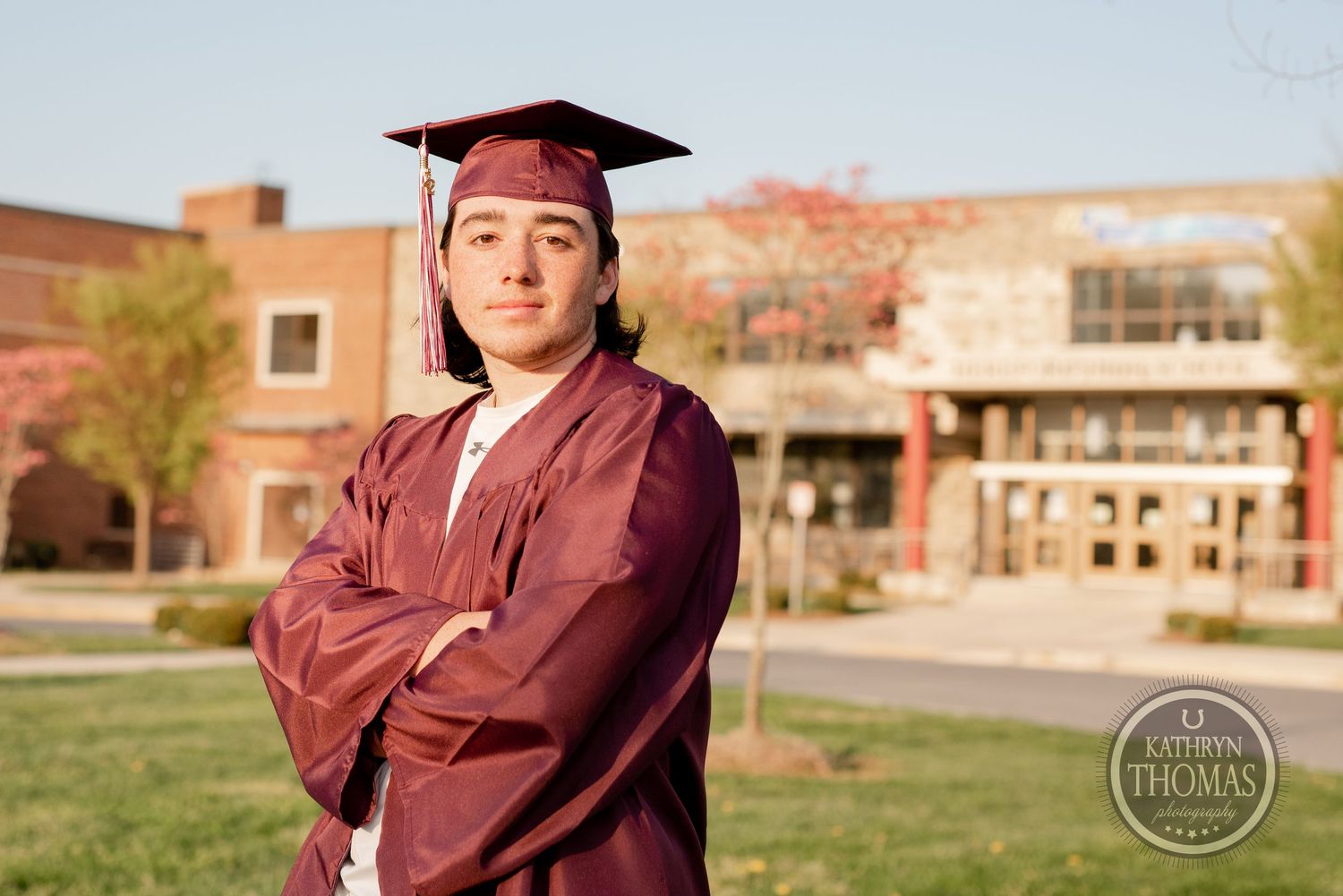 Graduate in maroon regalia stands confidently in front of brick university building.
