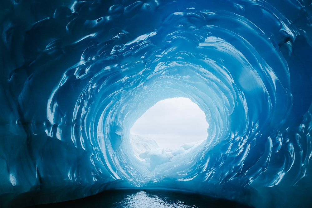 Ice cave interior with glowing blue walls and a bright opening to the sky.