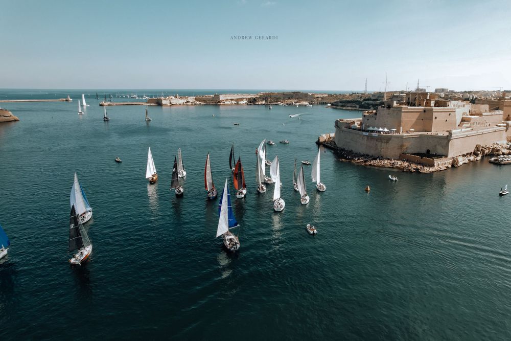 Aerial view of sailboats racing across blue Mediterranean waters near a coastal city.