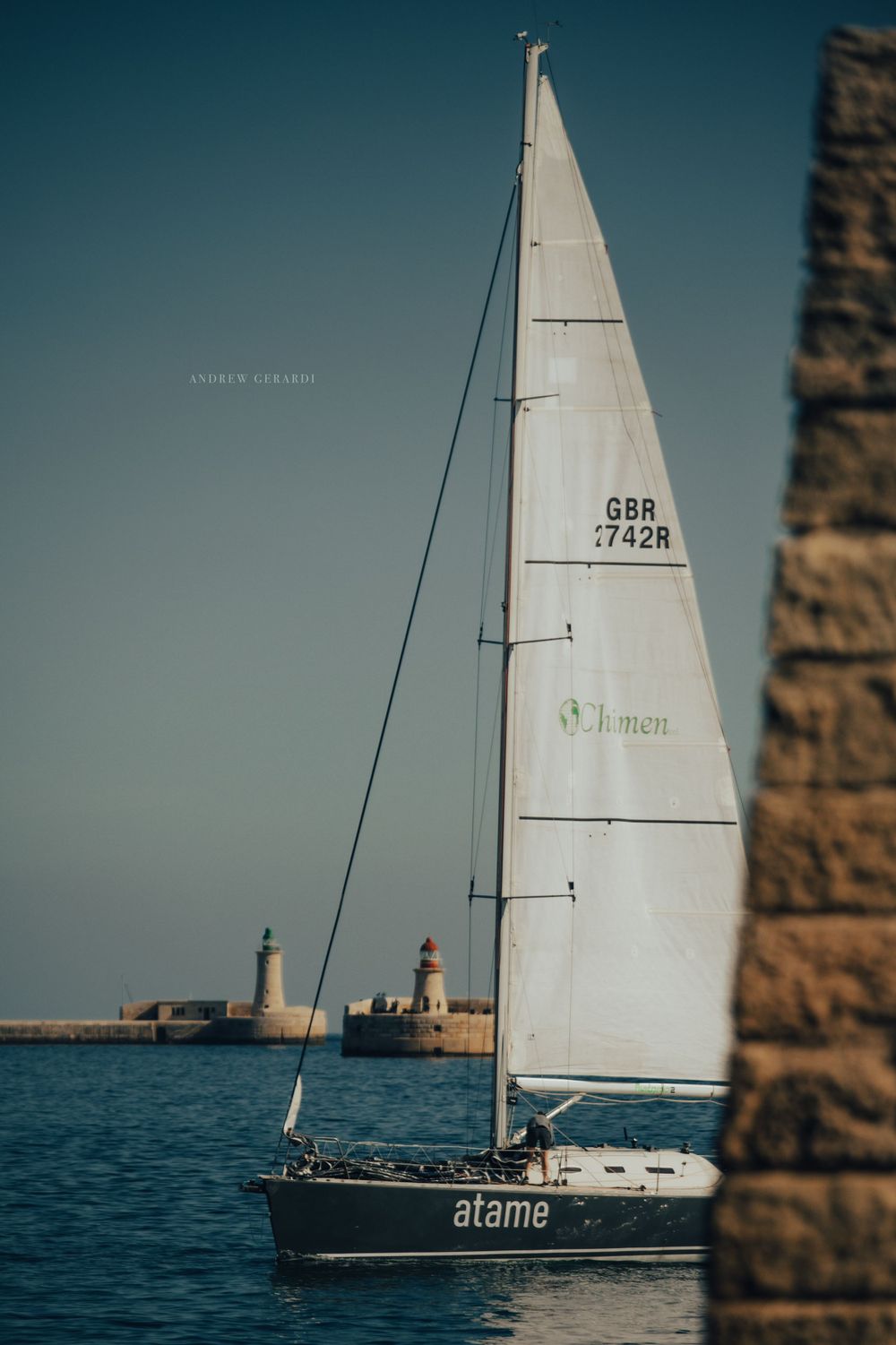 Sailboat named 'Atame' gliding through blue waters near a stone structure.
