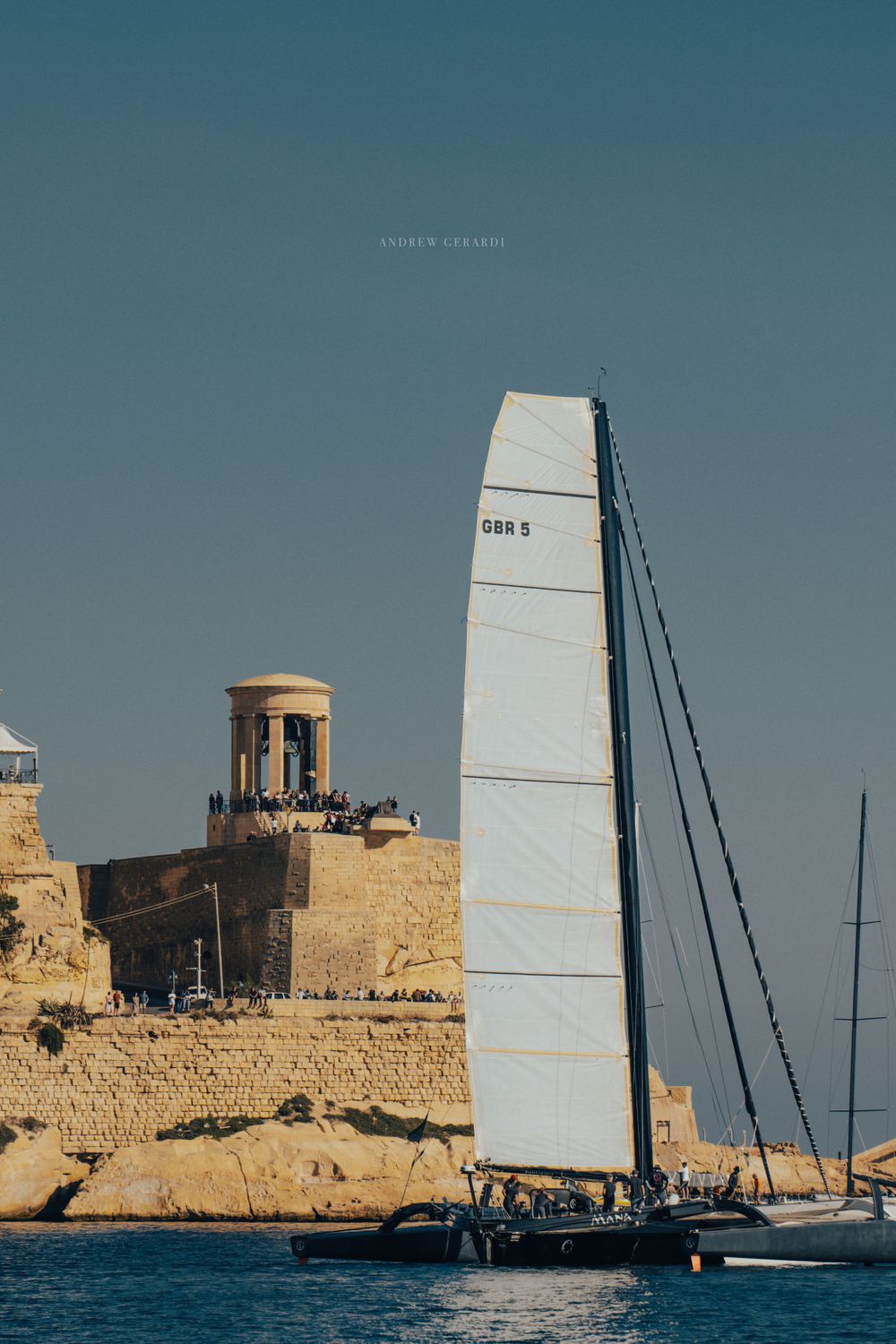 Large white sail of a racing yacht contrasts against historic stone fortifications along the coast.