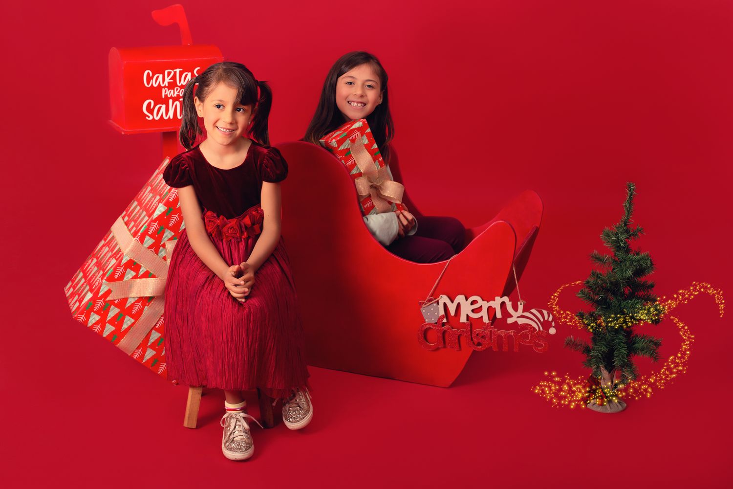 Two girls pose with a red Christmas sleigh prop and gifts against a vibrant red holiday backdrop.