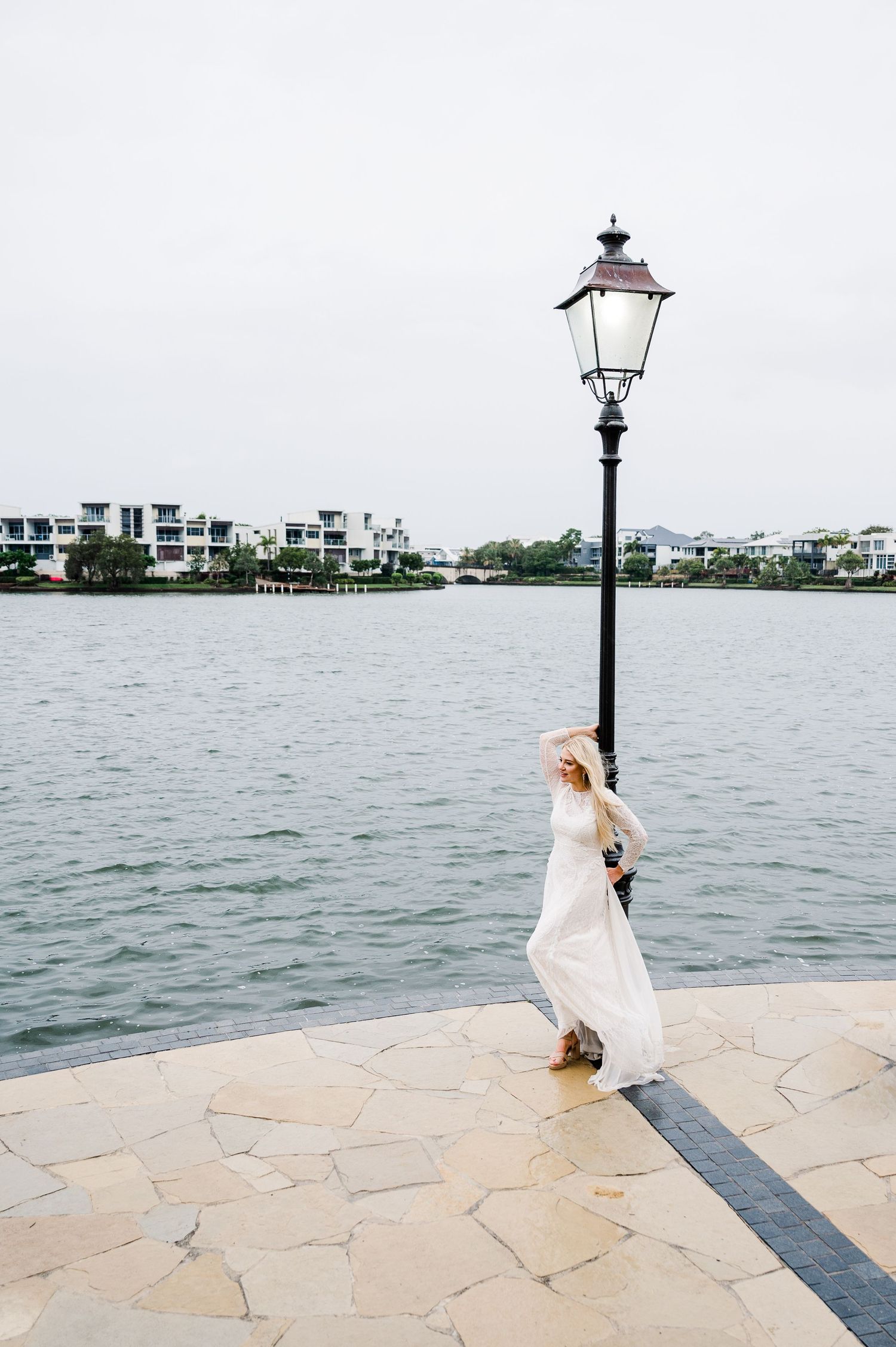 A figure in a flowing white dress poses dramatically by a waterfront lamppost.