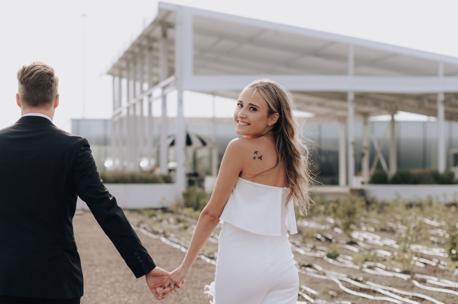 Bride in sleek white dress looks back while holding groom's hand.