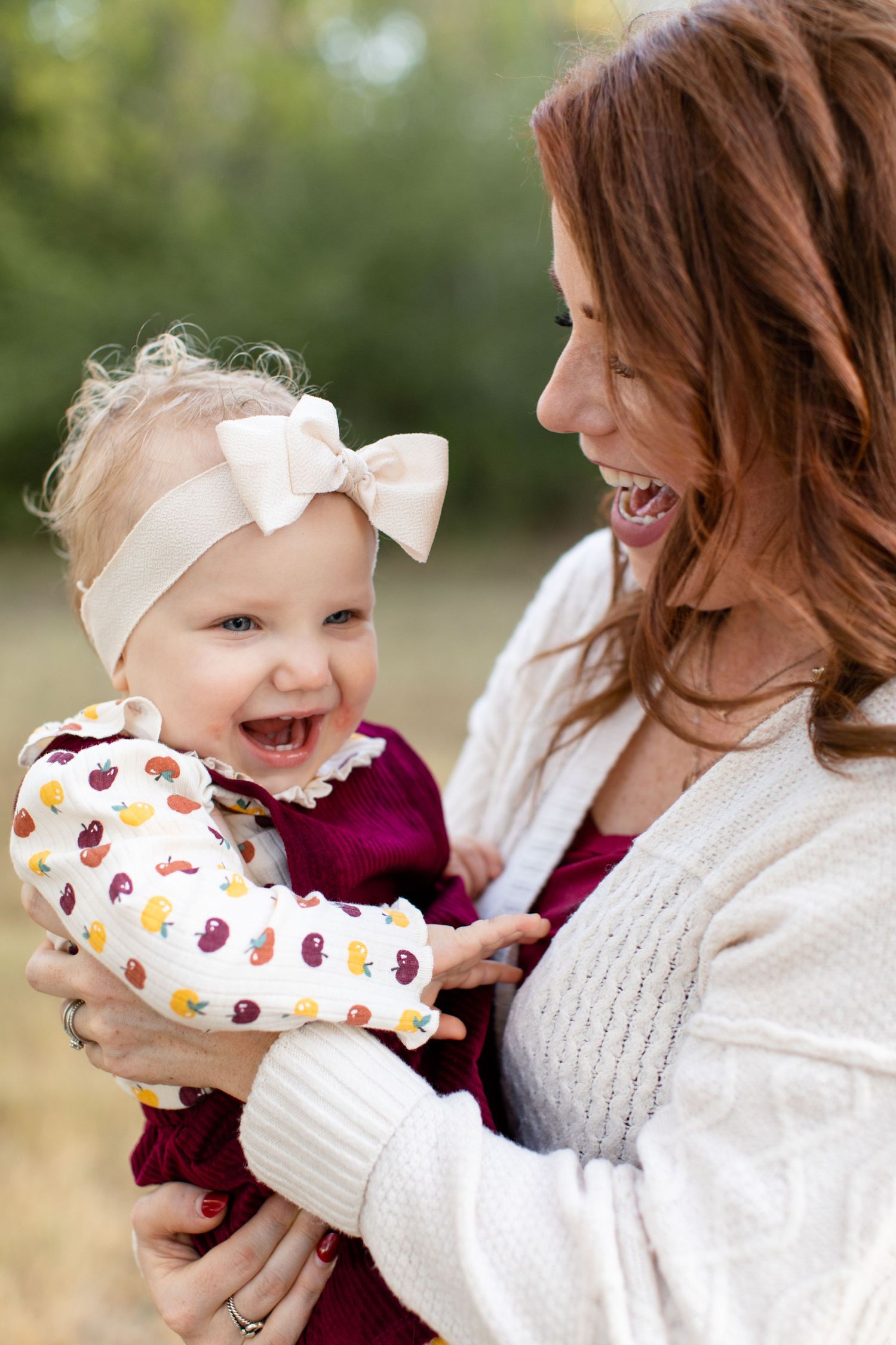 A happy baby in burgundy dress and bow headband shares laughter with their caregiver in white sweater.
