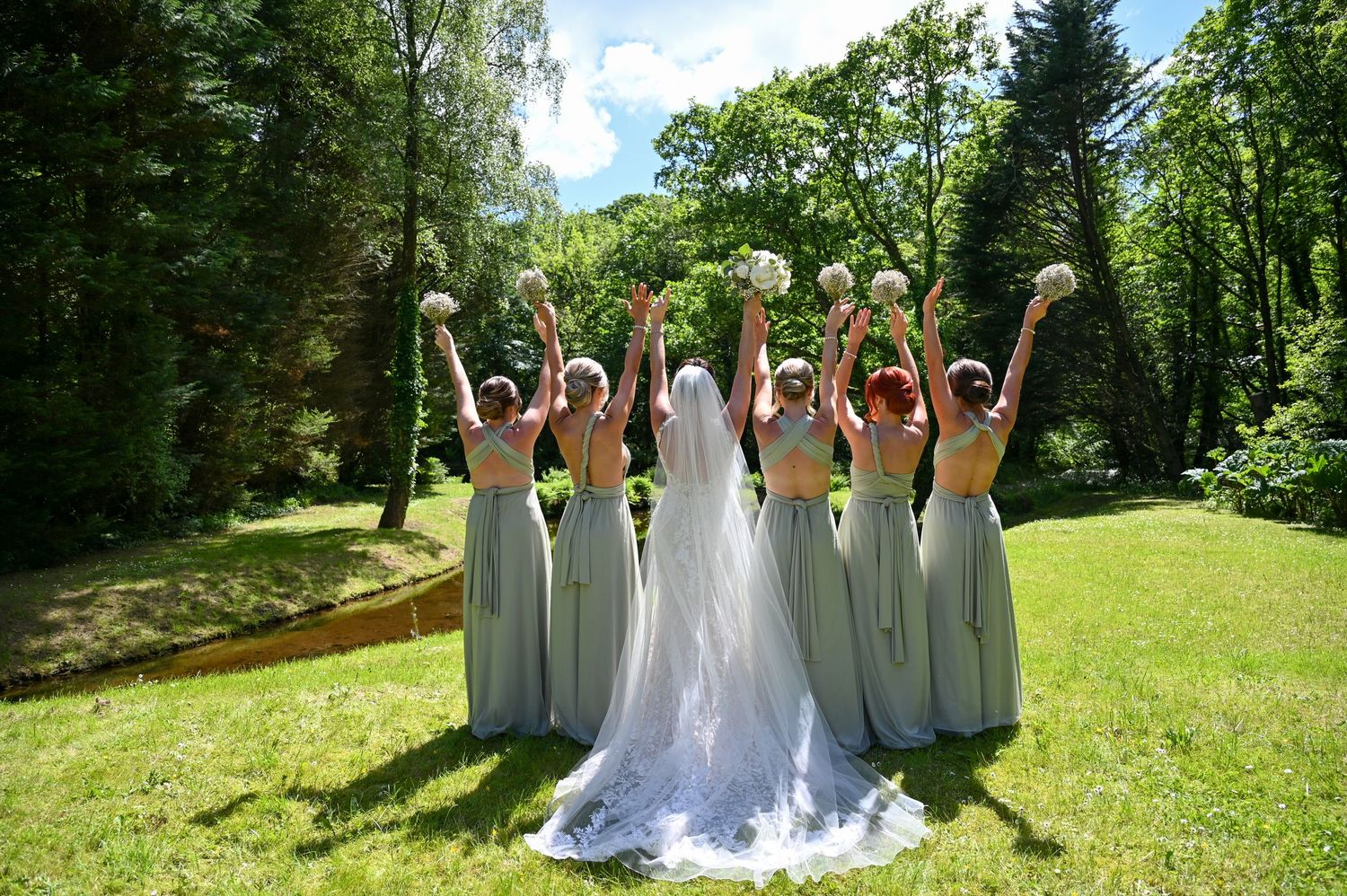 A bridal party in matching sage green dresses raise their bouquets together in a sunny garden setting.