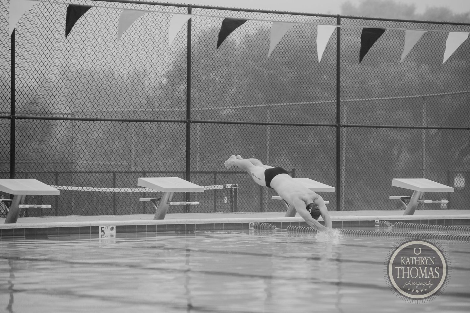 A swimmer diving off a starting block into a competition pool with pennant flags visible above.