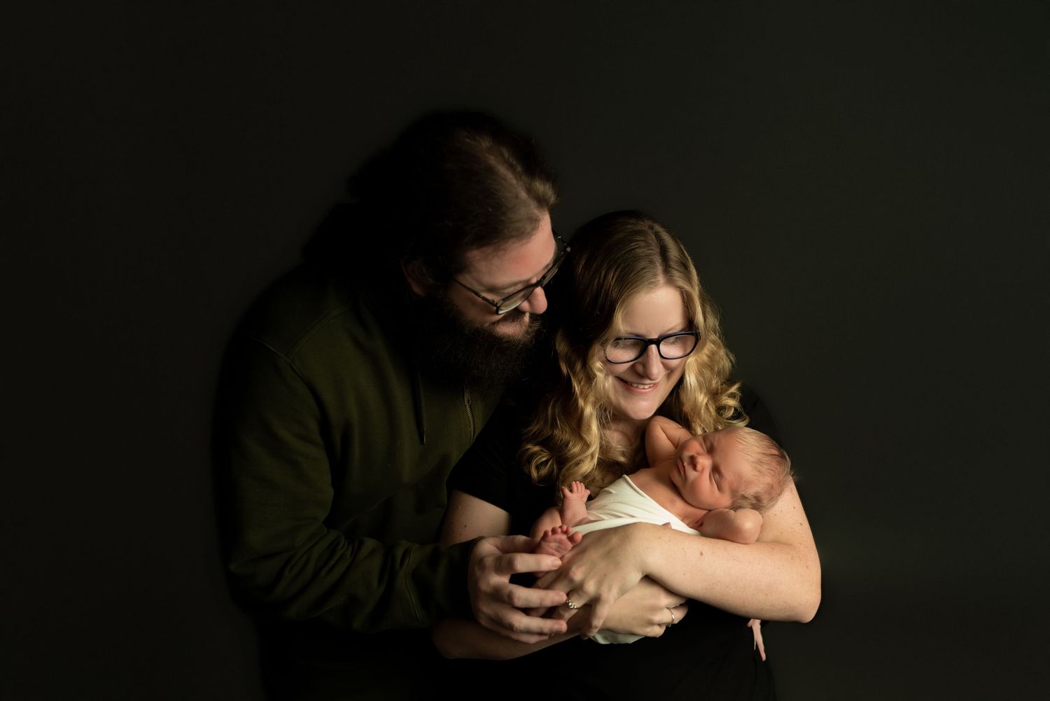 Intimate low-key studio portrait of a new family against dark background.