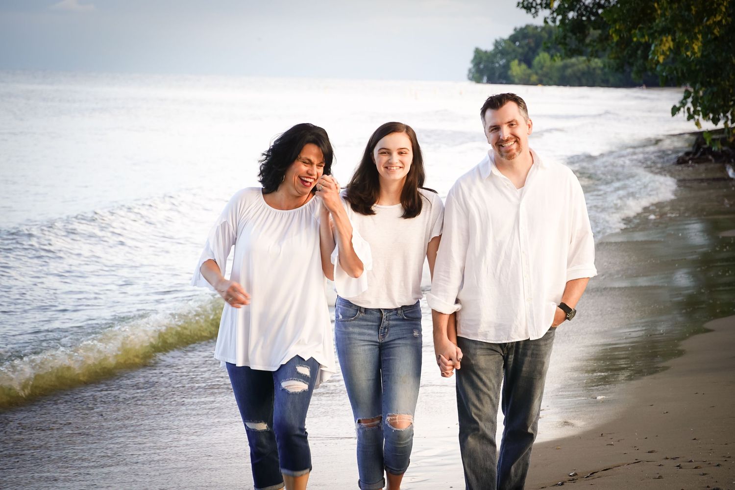 Three people in casual white outfits walking together along a peaceful beach shoreline.