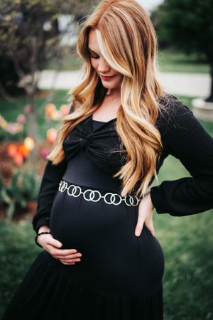 Expectant mother in black dress with chain belt poses in garden setting.
