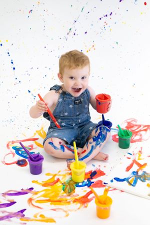 Toddler in denim overalls finger painting with colorful paints on white background.
