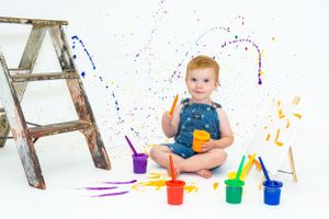 Baby in overalls plays with colorful paints next to wooden ladder against white backdrop.
