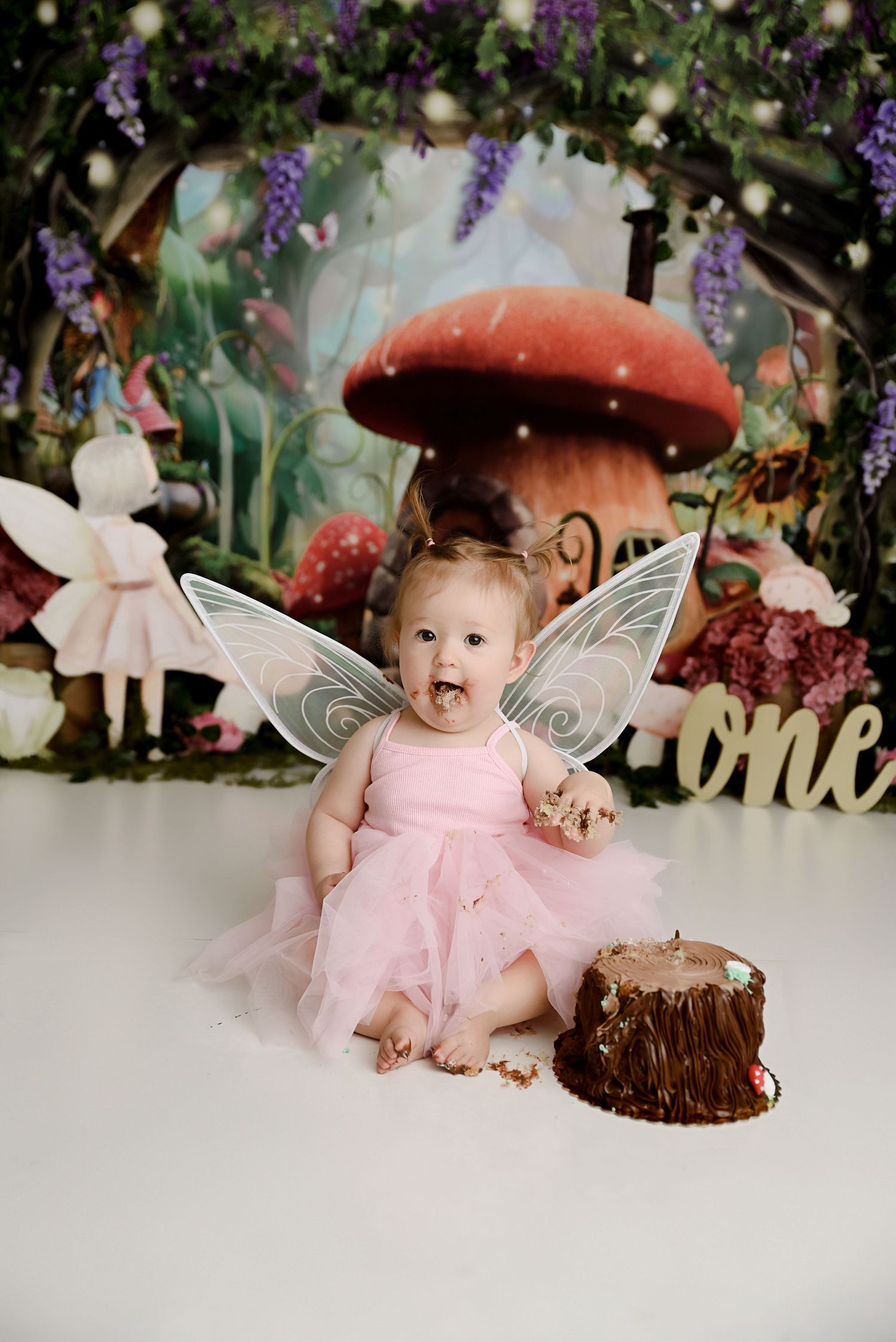 Baby girl in pink fairy costume with wings enjoys cake smash in enchanted mushroom forest setting.