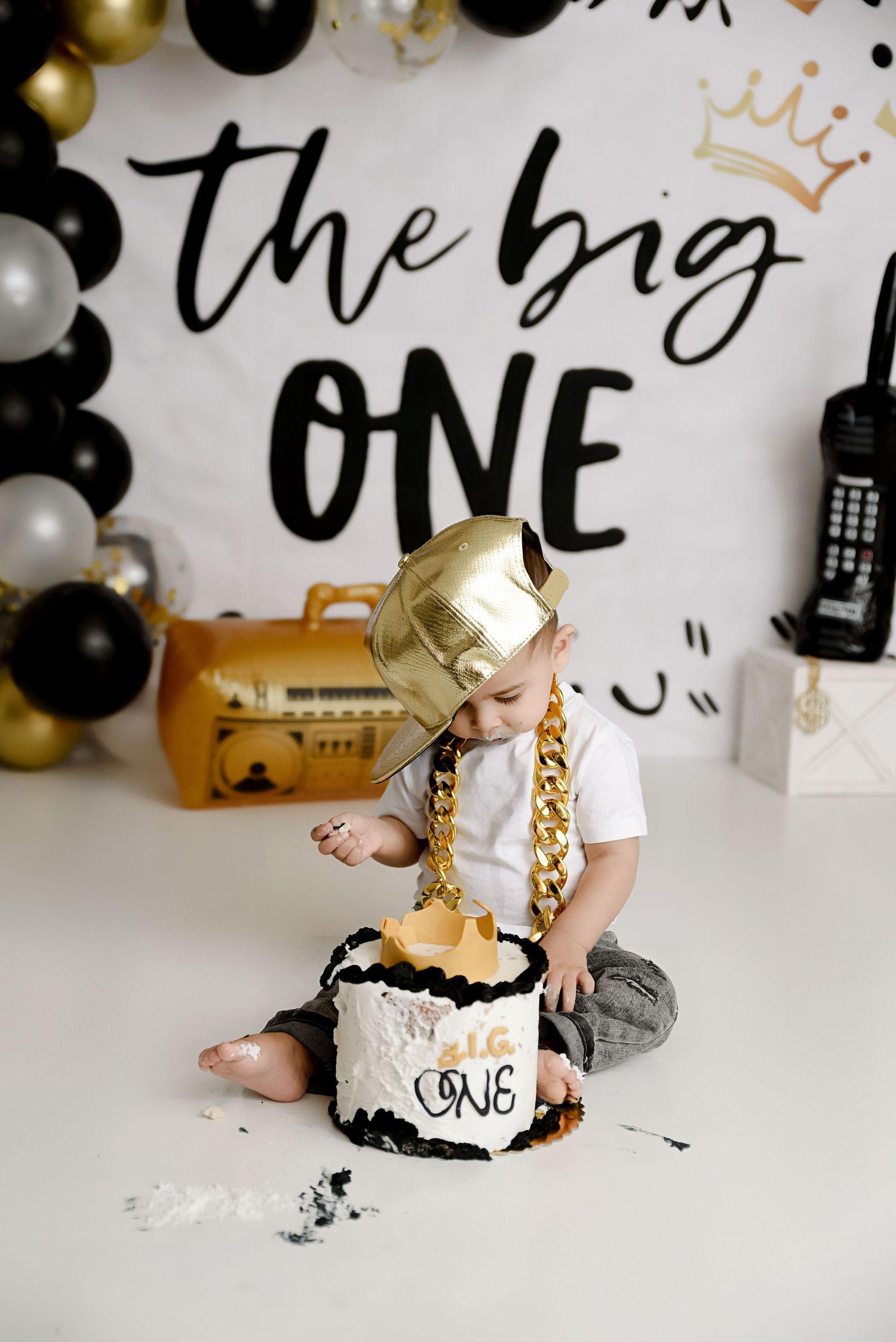 Baby boy in gold chain and hat smashes first birthday cake at a hip-hop themed cake smash session.