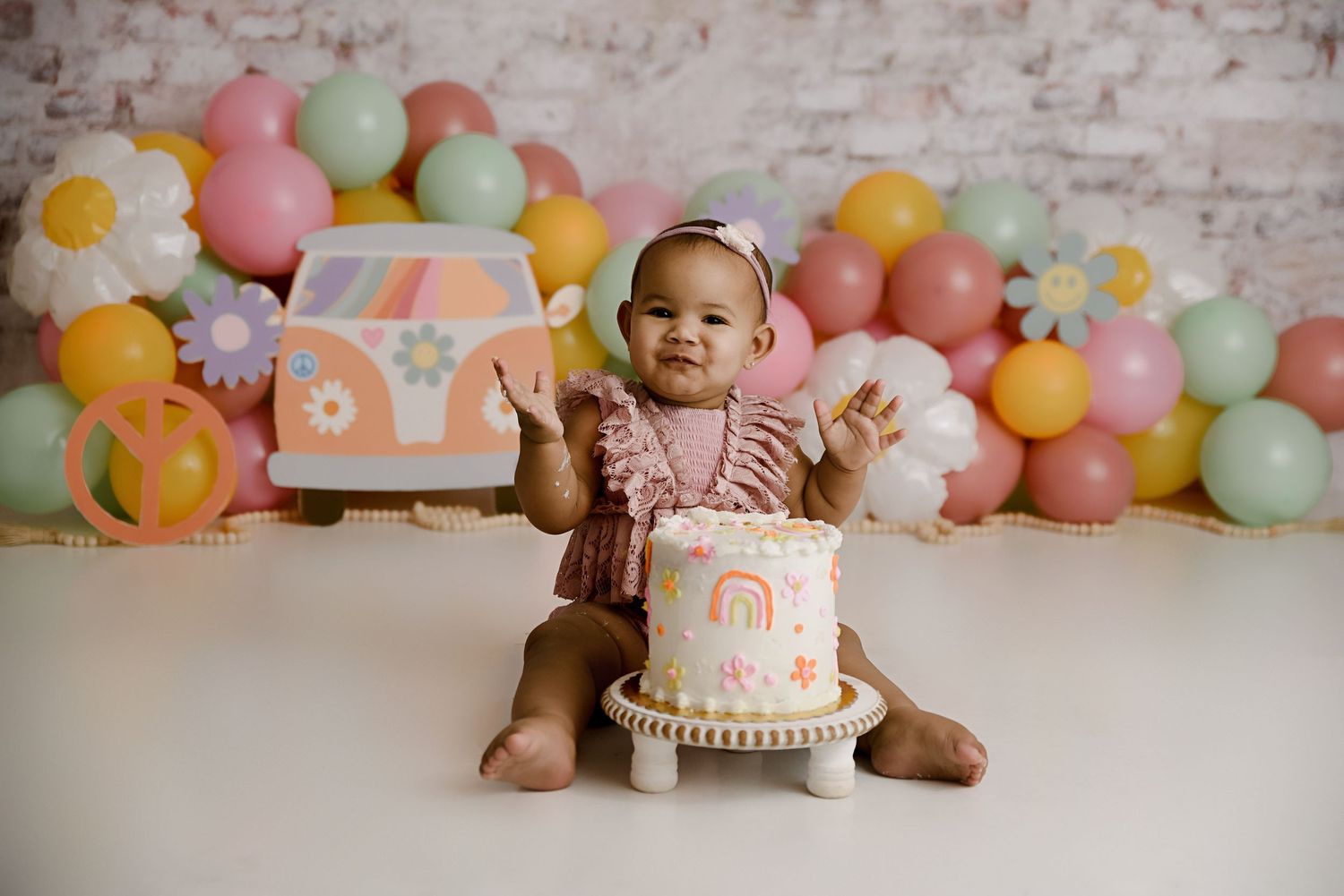 Smiling baby girl in pink romper claps hands beside colorful retro boho cake smash setup with balloons.