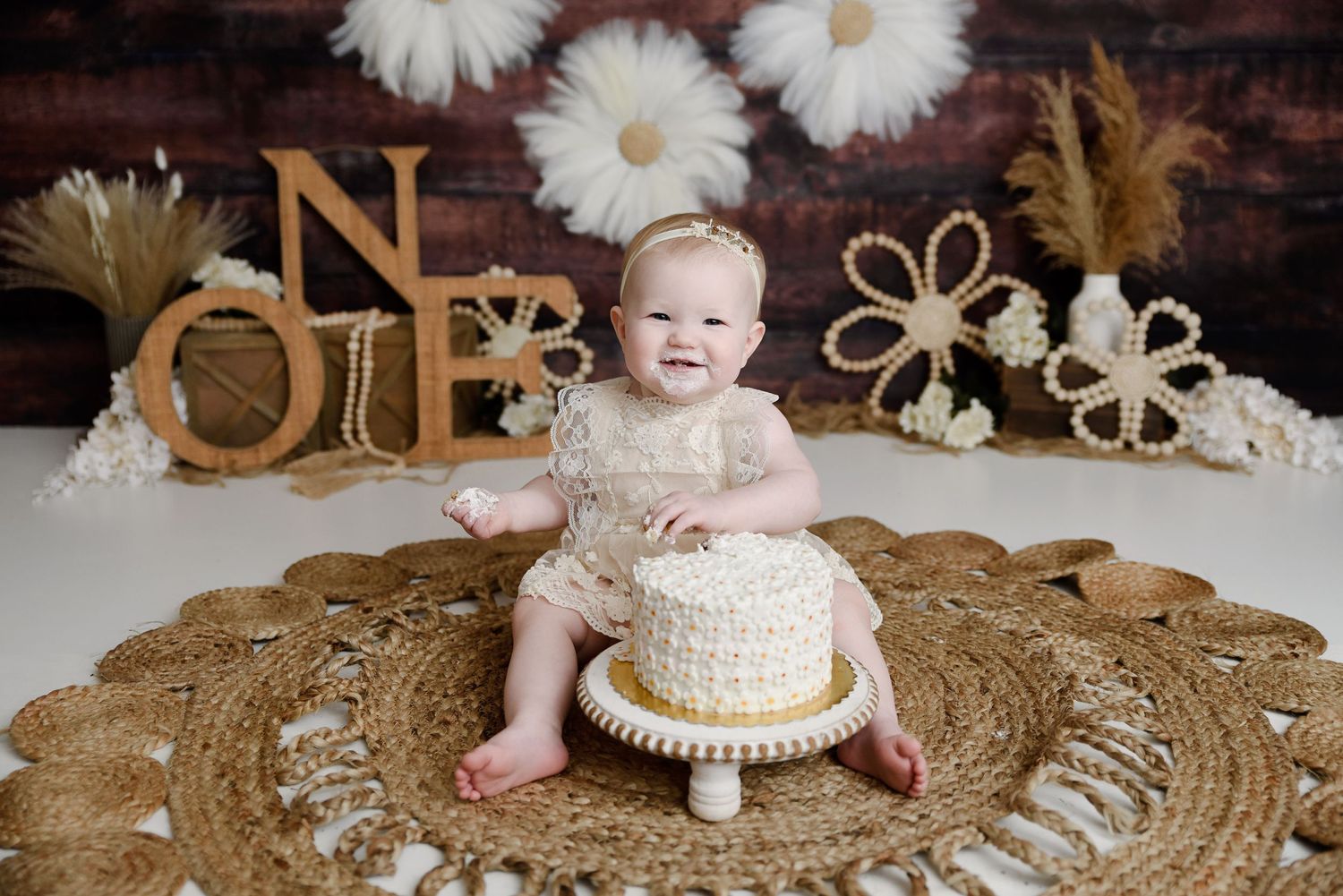 Baby girl smiling at boho-themed first birthday cake smash on jute rug with floral decorations.