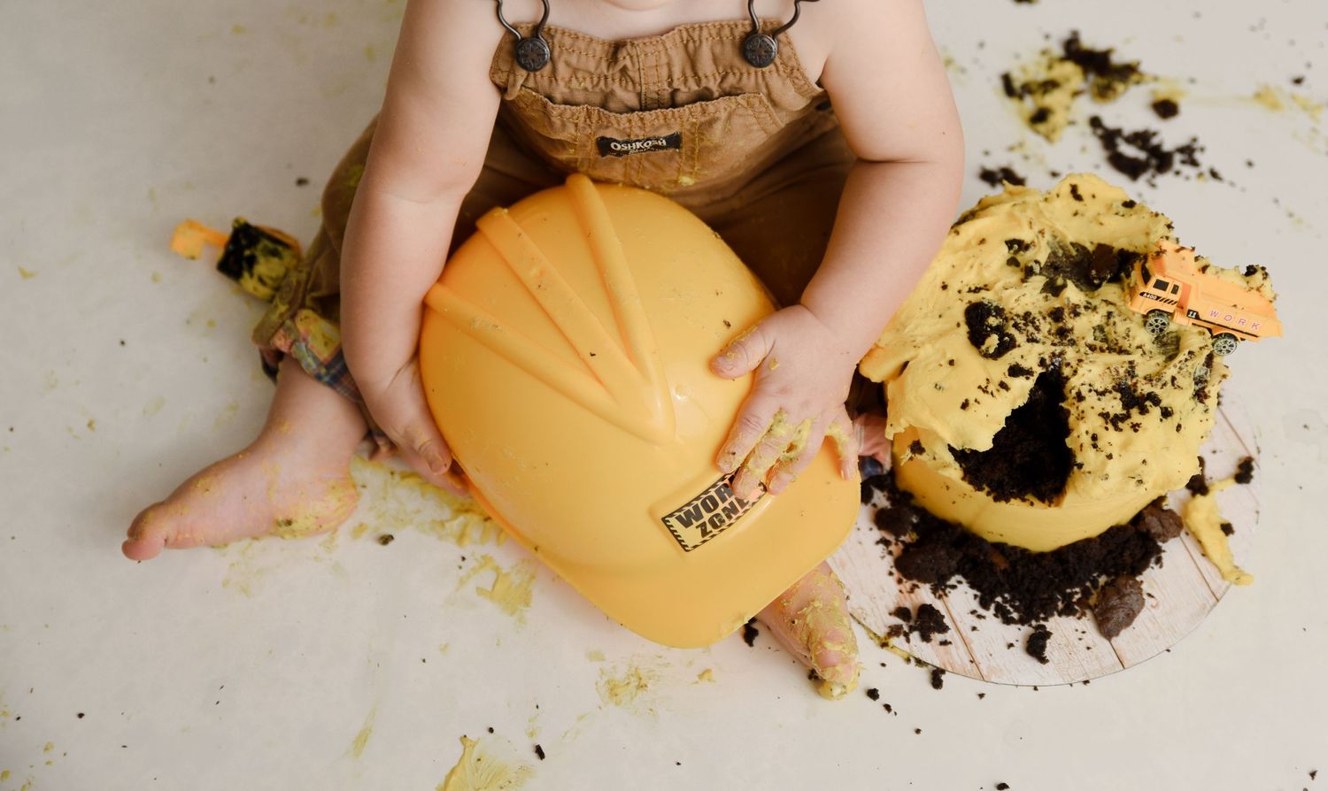 Baby in overalls holds yellow hard hat during construction-themed first birthday cake smash session.
