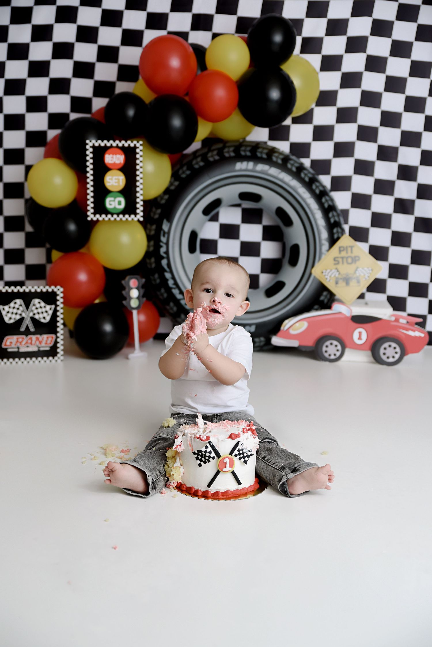 Baby boy claps hands beside race car themed first birthday smash cake with checkered flag backdrop.