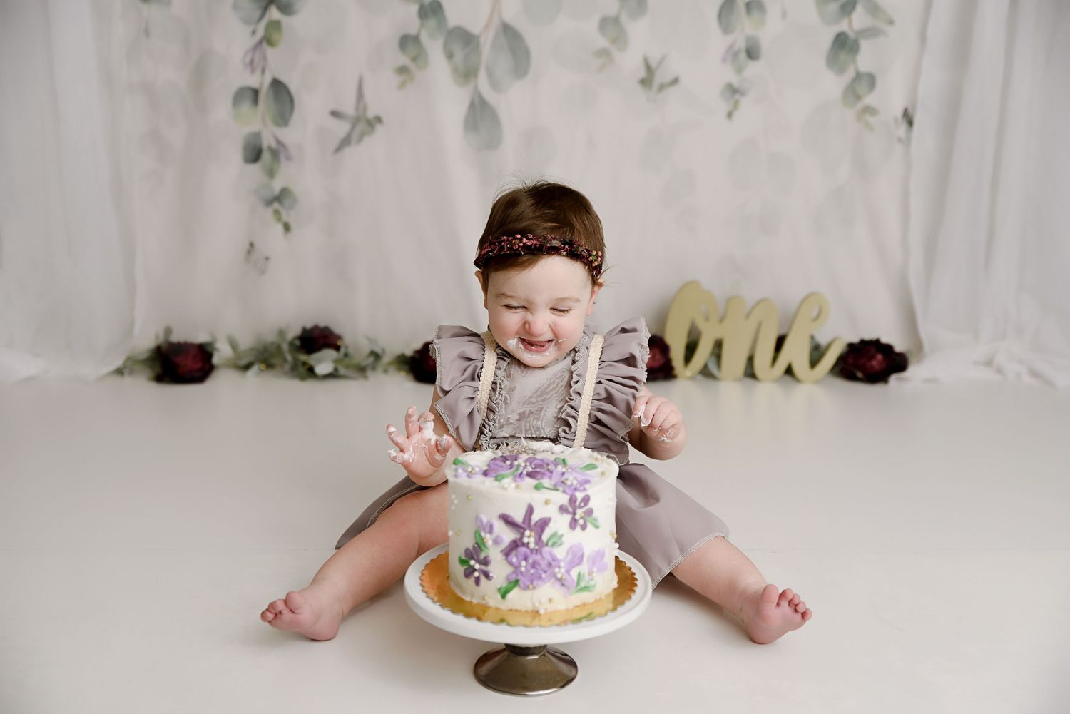 Baby girl in grey dress laughs during floral purple cake smash with greenery backdrop and one sign.