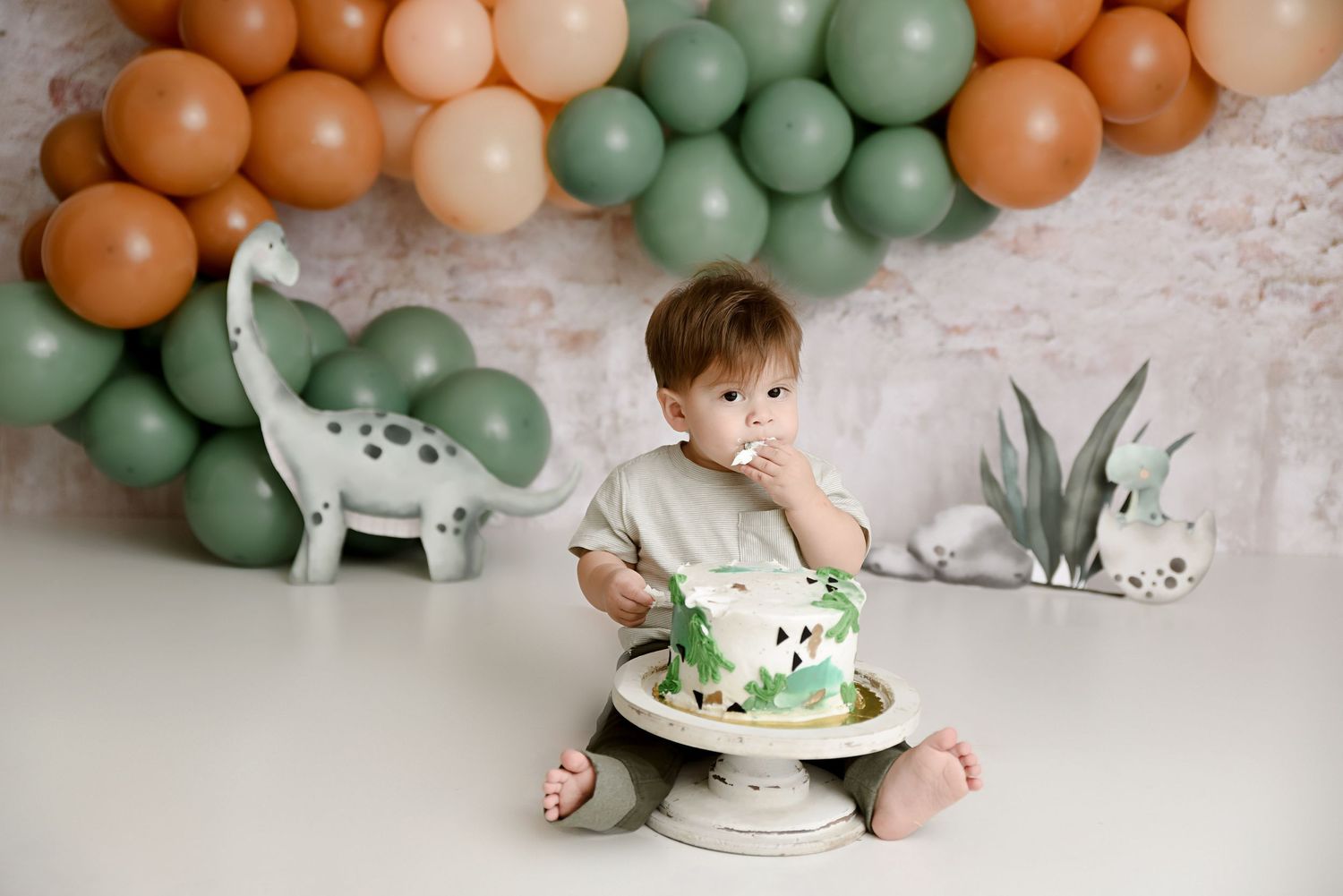 Toddler boy enjoys dinosaur-themed first birthday cake smash with green and orange balloon garland.