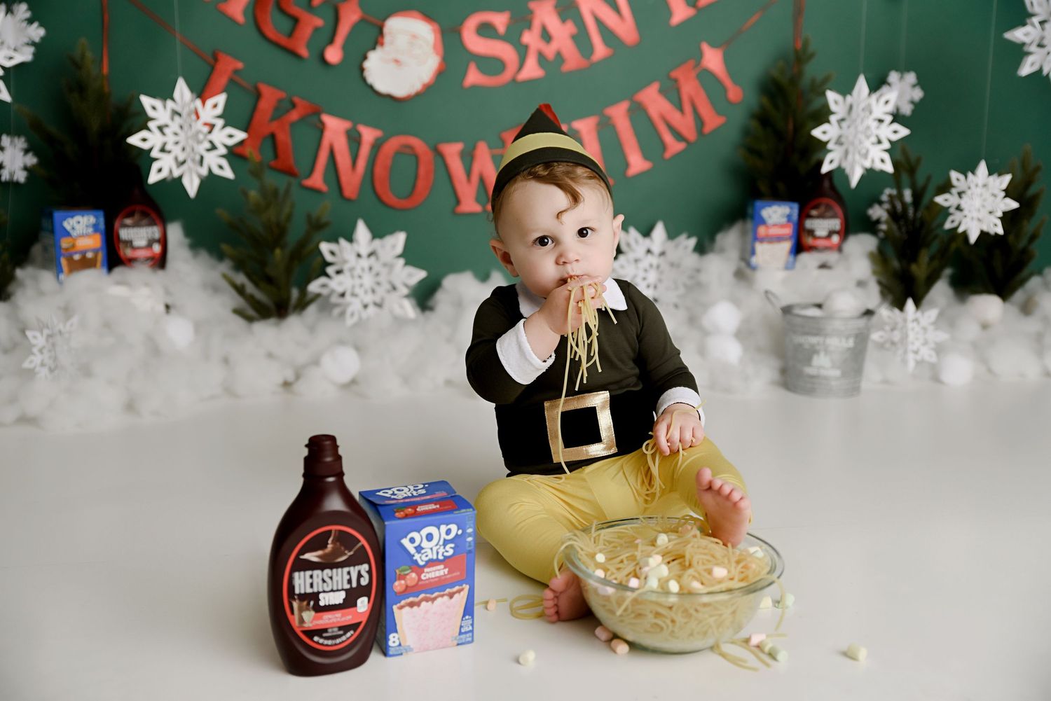 Baby dressed as an elf enjoys a Christmas-themed cake smash with Hershey's syrup and popcorn.