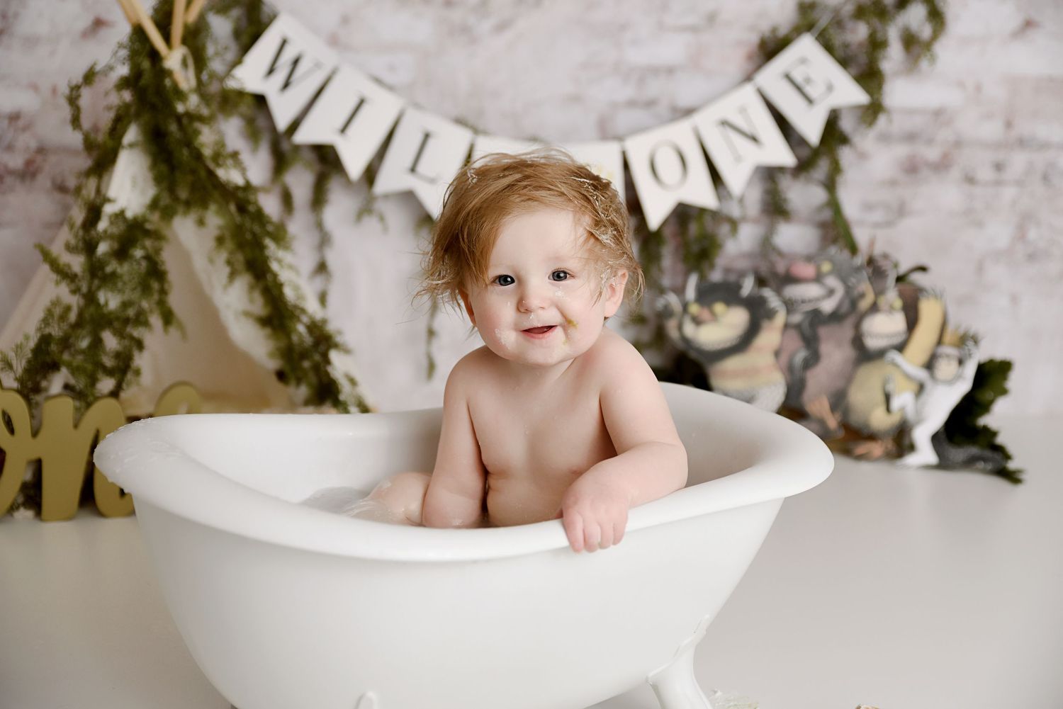 Smiling red-haired toddler sits in white bathtub during Wild One first birthday bath splash session.